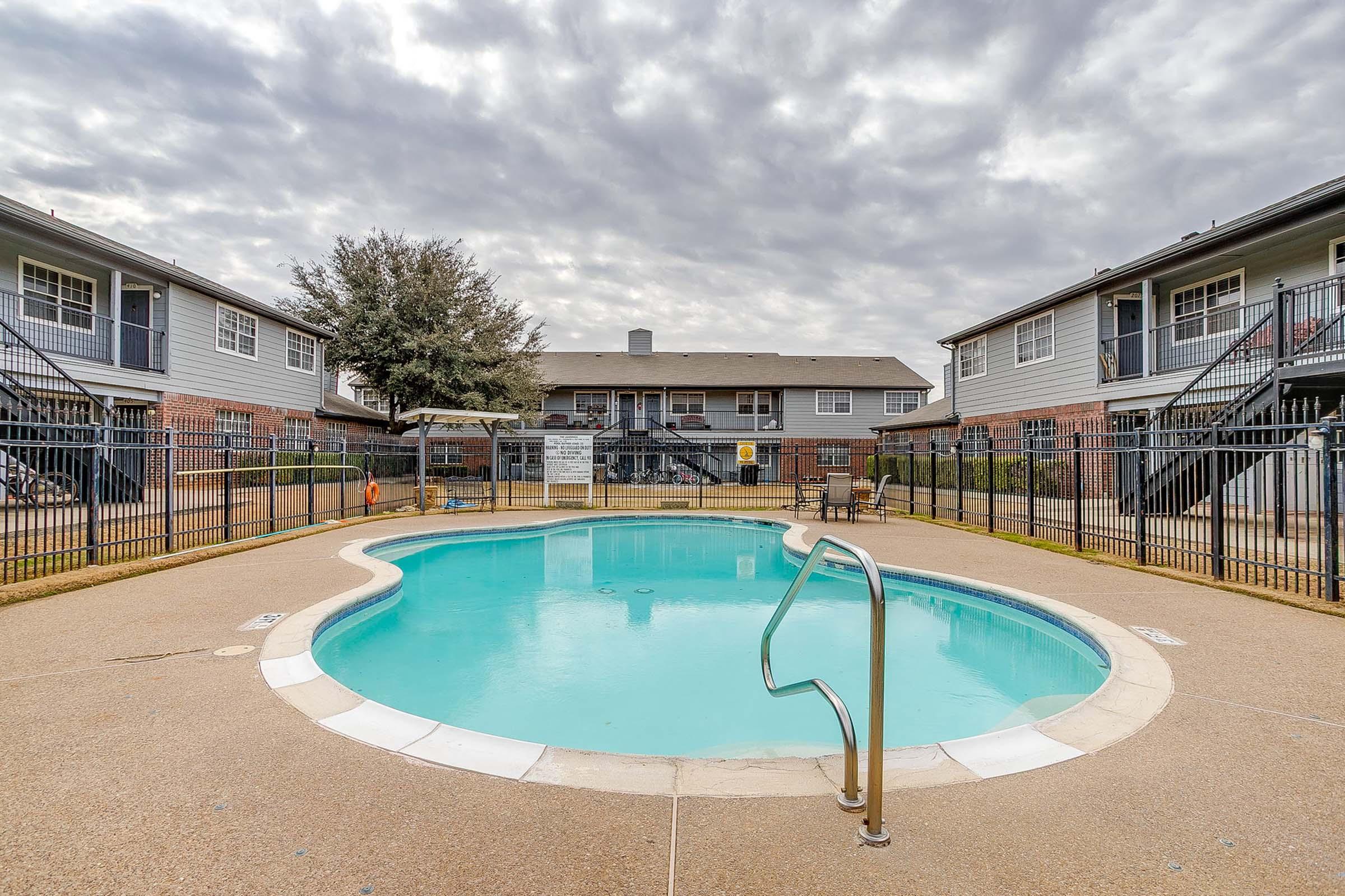 A clear blue swimming pool surrounded by a light-colored, textured deck. The pool is enclosed with a black metal fence. In the background, there are two-story apartment buildings with balconies on either side, and a cloudy sky overhead. Some lounge chairs are positioned around the pool area.
