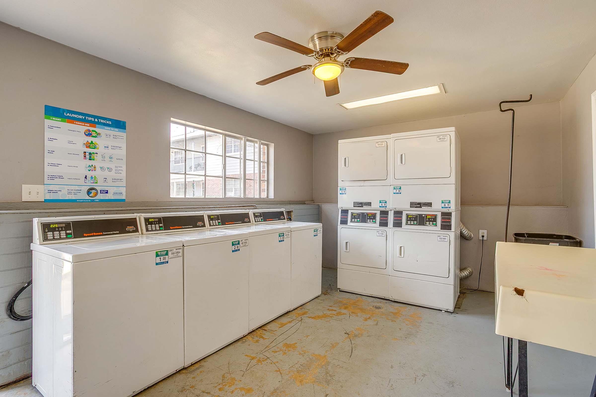 Interior of a laundry room featuring several white washing machines and dryers lined against the wall. A ceiling fan is visible, and a window allows natural light to enter. A utility sink is in the corner, and an informational poster about laundry operations is displayed on the wall.