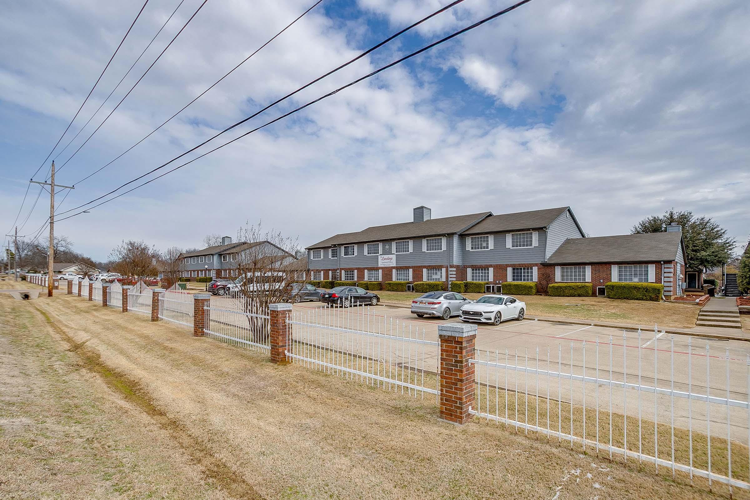 A view of a residential apartment complex with a paved parking area, featuring multiple buildings, a well-maintained lawn, and a white fence. The sky is partly cloudy, and there are utility poles and trees in the background, creating a suburban atmosphere.