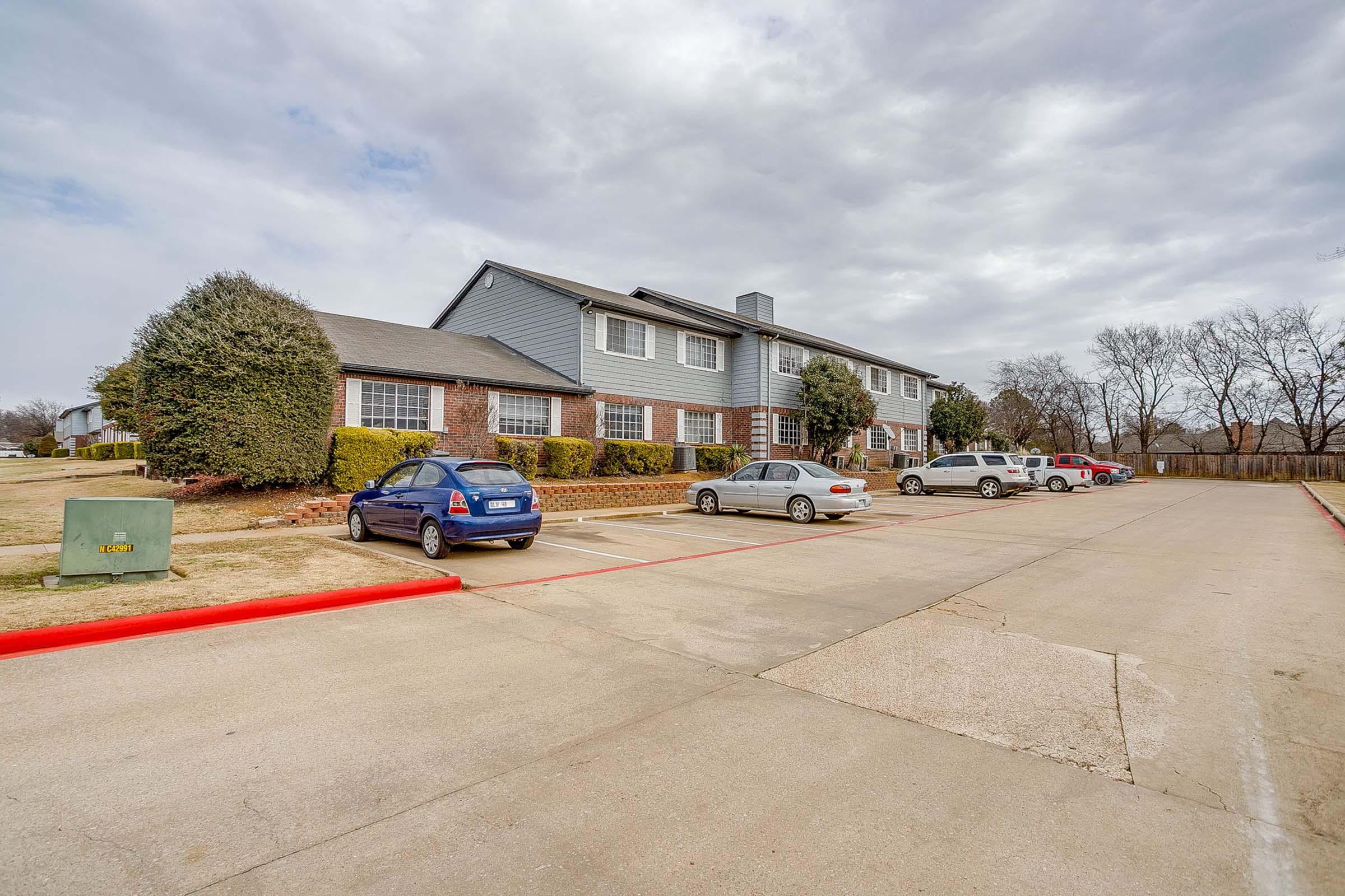 A multi-story building with a brick facade and gray siding, surrounded by neatly trimmed shrubs. Several parked cars are visible in a parking lot in front of the building, and a cloudy sky looms overhead. The area appears well-maintained and suburban in setting.