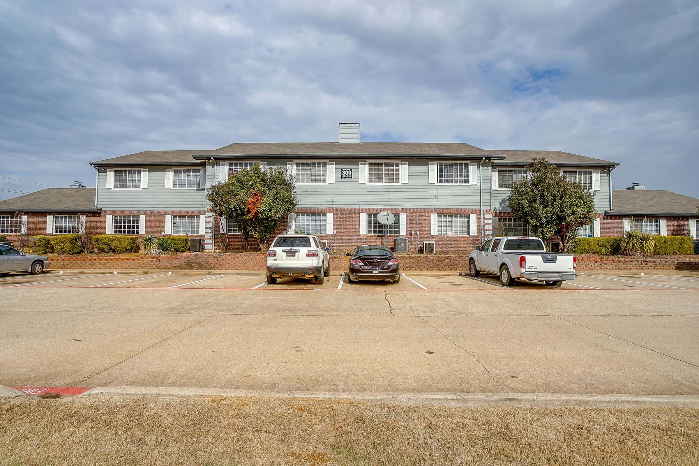 A two-story brick apartment building with multiple windows and a light green section. There are several parked cars in front, including a white SUV, a black sedan, and another white vehicle. The parking lot is surrounded by grass and a few trees, under a cloudy sky.