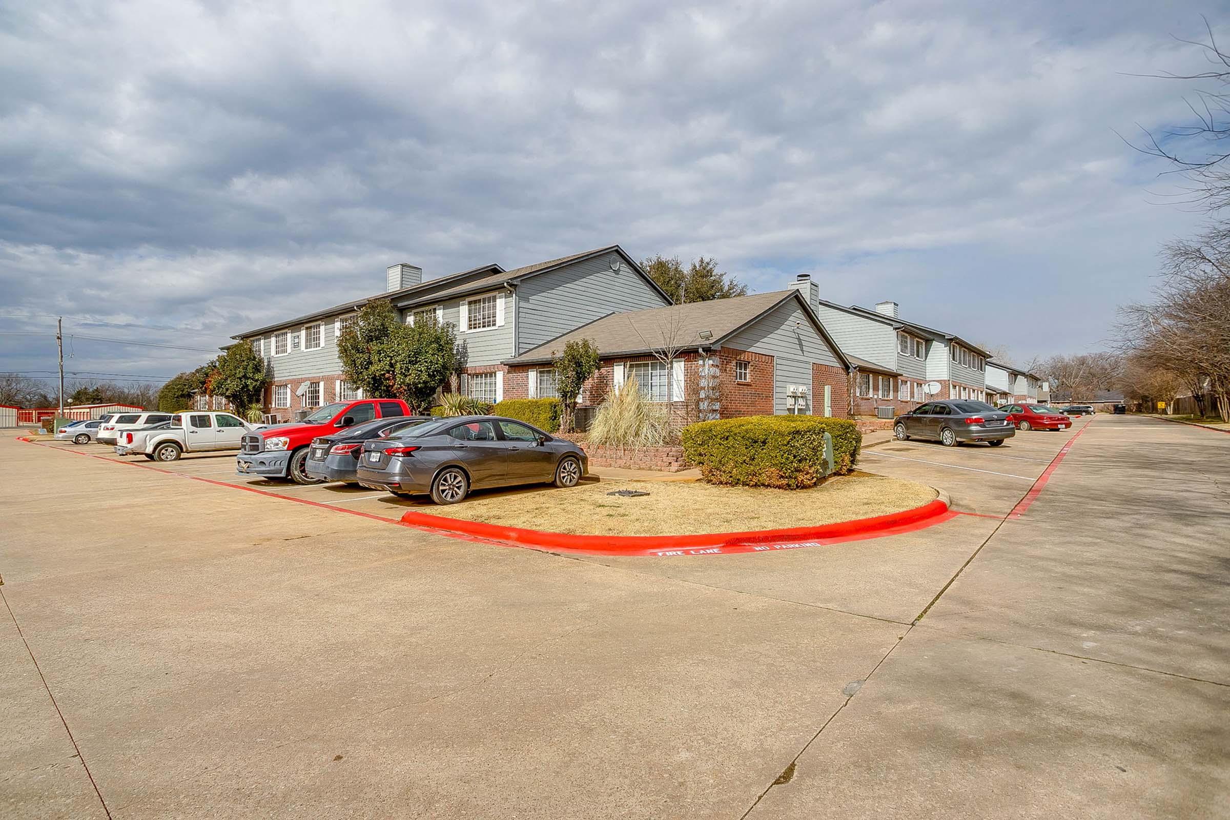 A quiet residential area featuring several two-story apartment buildings with brick exteriors. Cars are parked in a well-maintained lot, and the surrounding landscape includes some greenery and bare trees under a cloudy sky. The overall scene conveys a calm and suburban atmosphere.