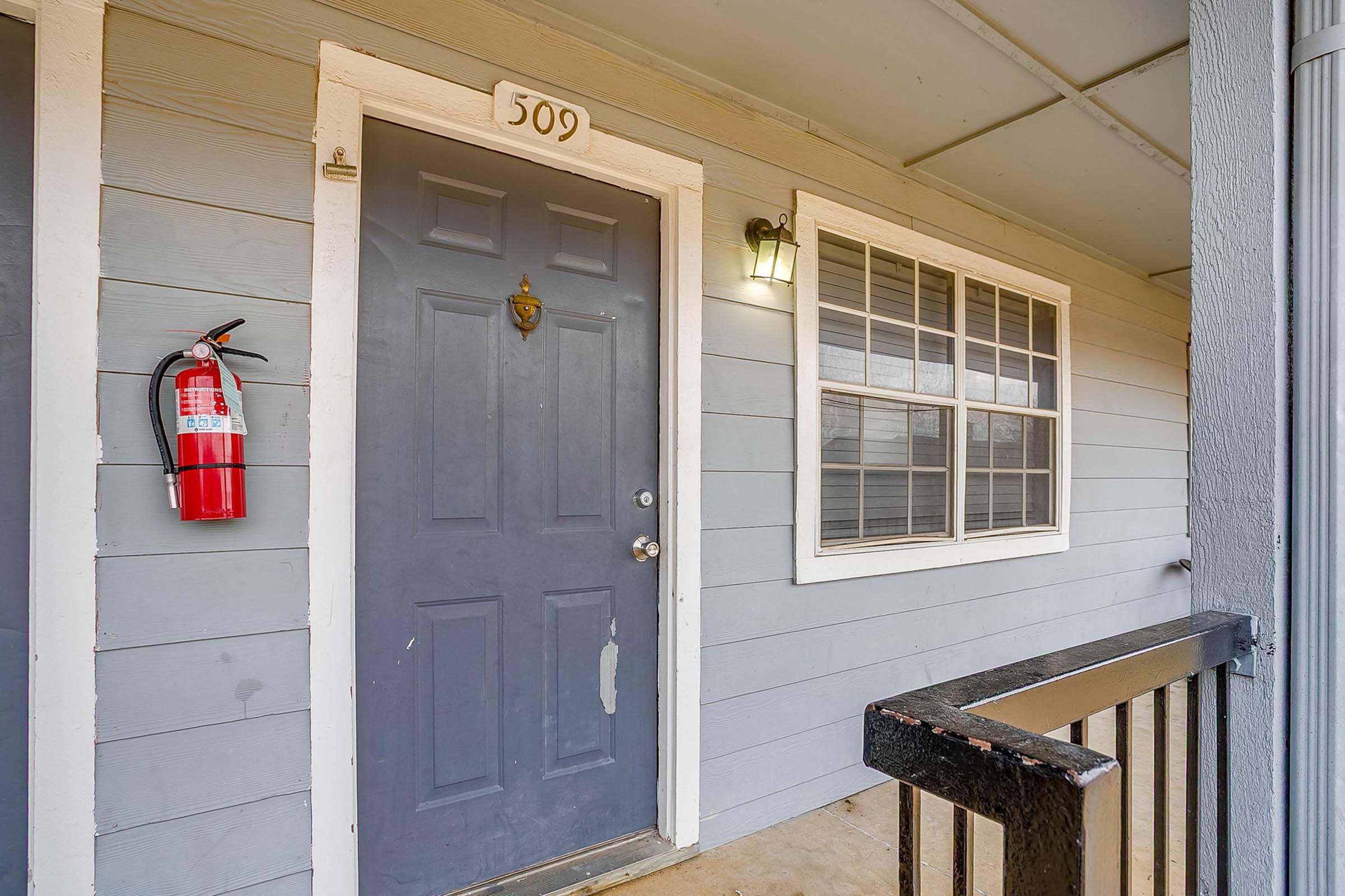 A blue exterior door with the number 509 displayed. There is a wall-mounted fire extinguisher to the left and a window with multiple panes to the right. The porch area features a railing, and the scene is well-lit.