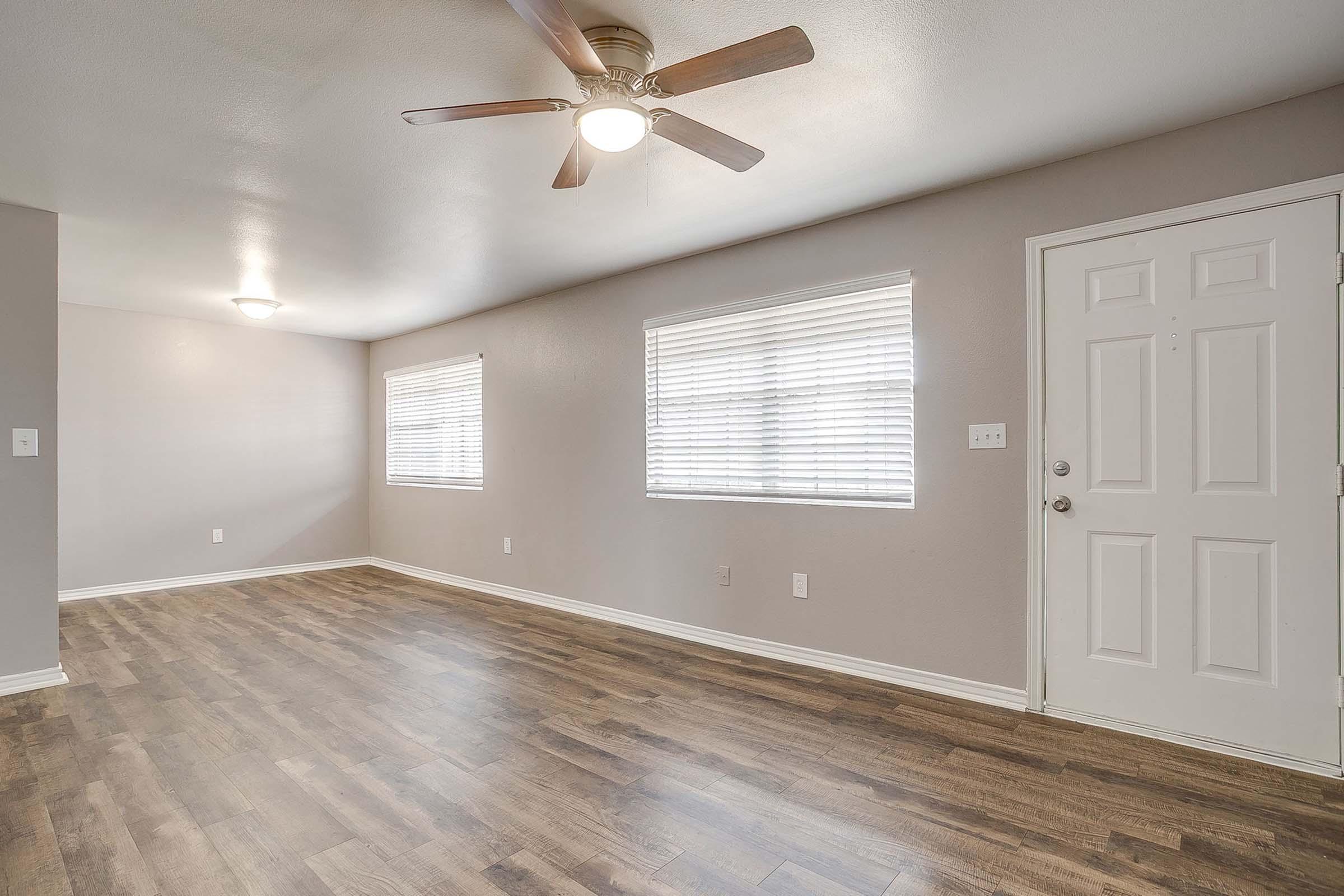 A spacious living room with light gray walls and wooden plank flooring. It features a ceiling fan, two windows with white blinds, and a front door. The room is well-lit and empty, creating a bright and inviting atmosphere.