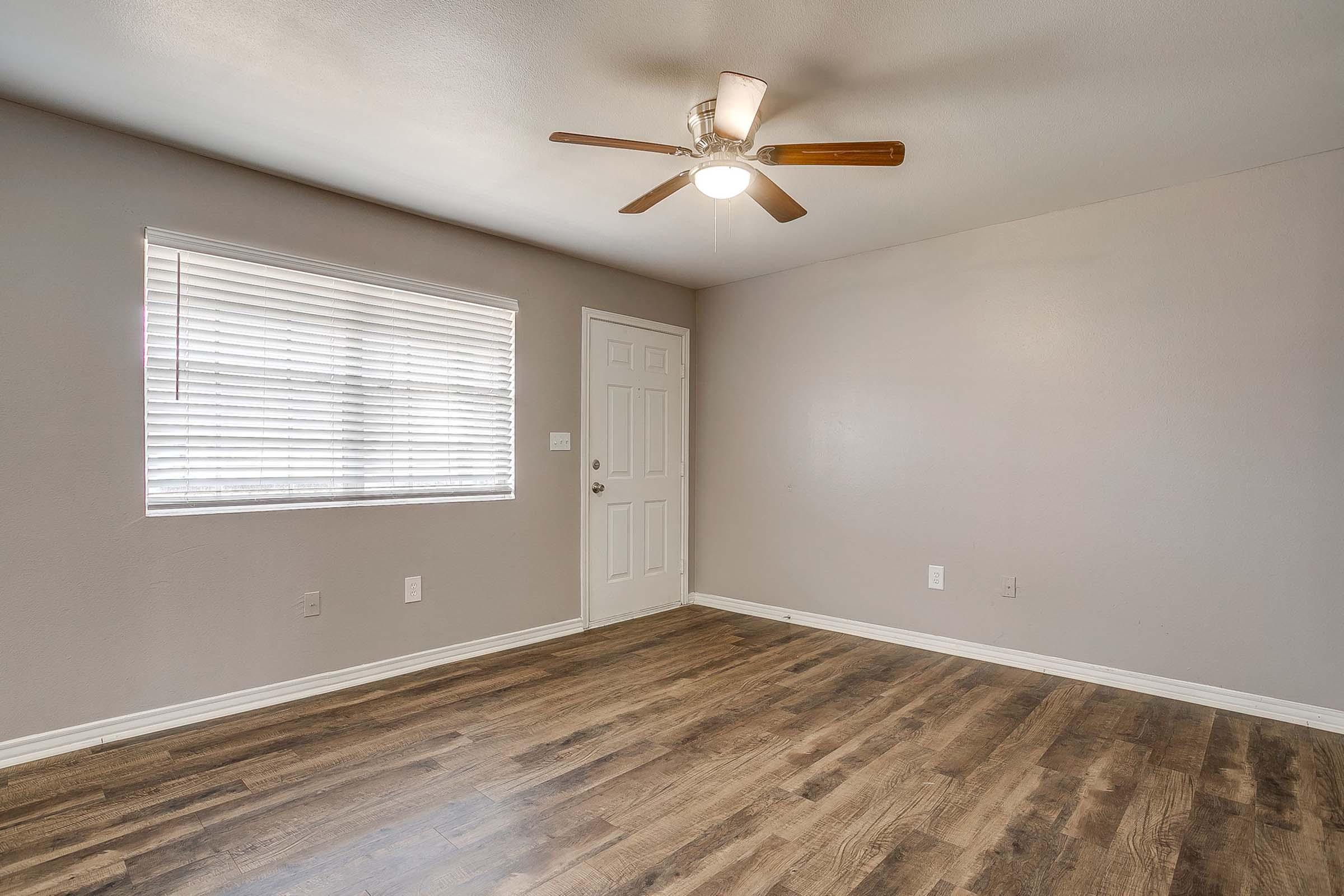 Empty room featuring a ceiling fan, a wooden floor, and a window with blinds. A plain door is visible on the right wall, and the walls are painted in a soft gray color. The space is well-lit, showcasing a minimalist and clean design suitable for various purposes.