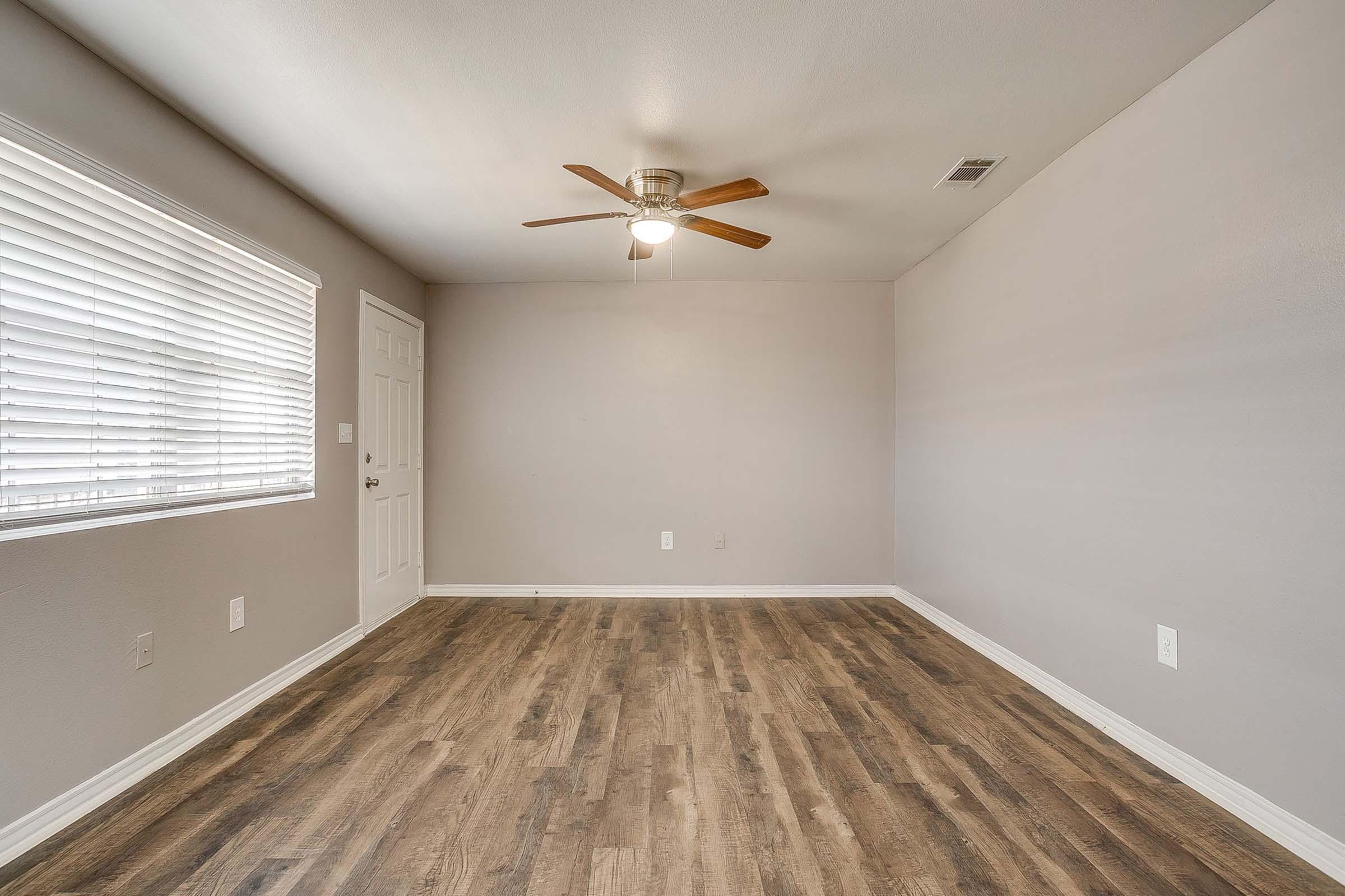 Empty room with light gray walls and hardwood-style flooring. A ceiling fan with wooden blades is centered in the room. A window with white blinds lets in natural light, and there's a door on the left side. The space appears clean and uncluttered, ready for furnishings.