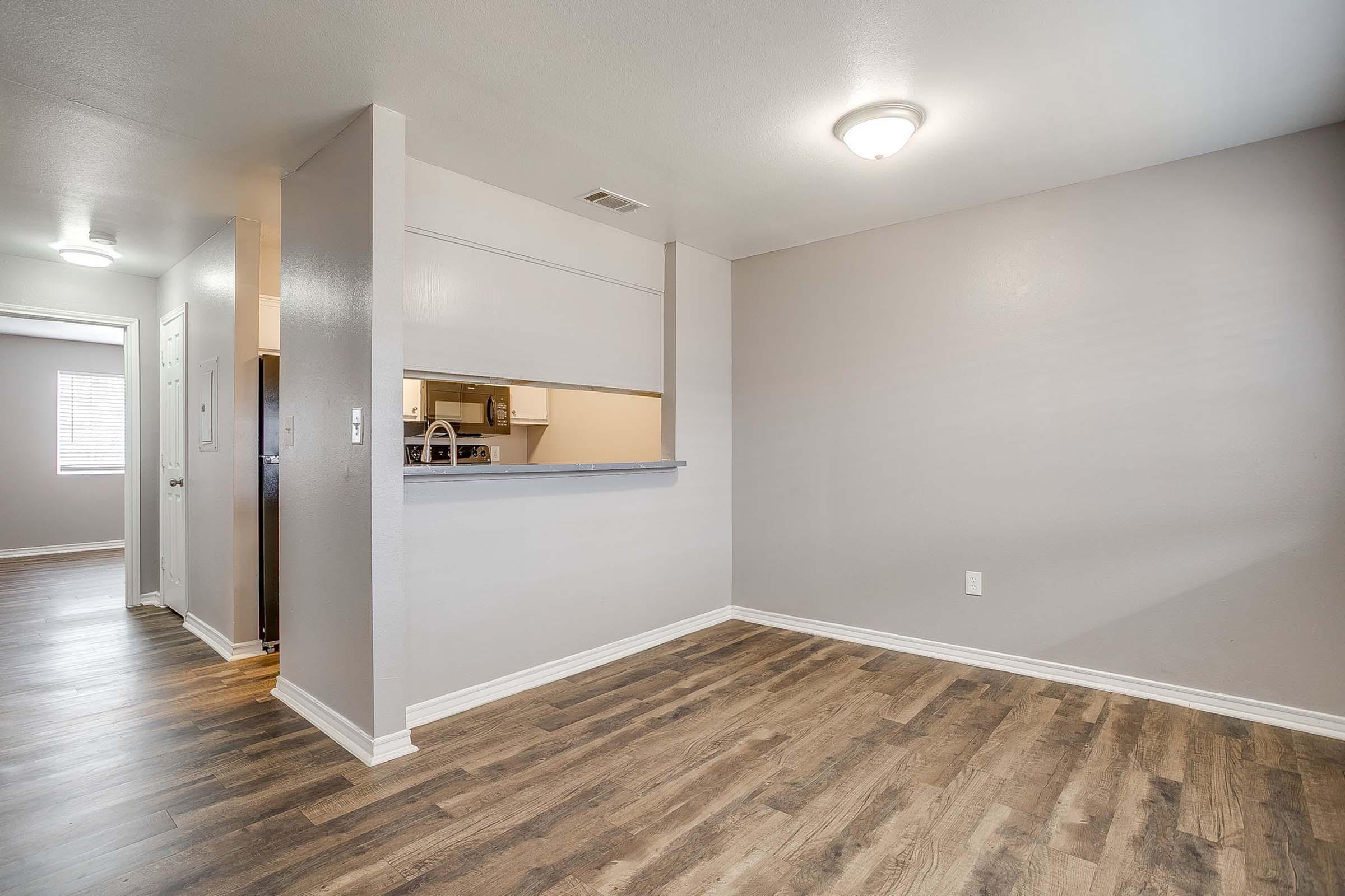 Spacious interior of a modern apartment featuring light gray walls, a wood-look laminate floor, and recessed lighting. An open layout connects the living area to a kitchen with a serving bar. Windows in the background provide natural light, creating a bright and inviting atmosphere.
