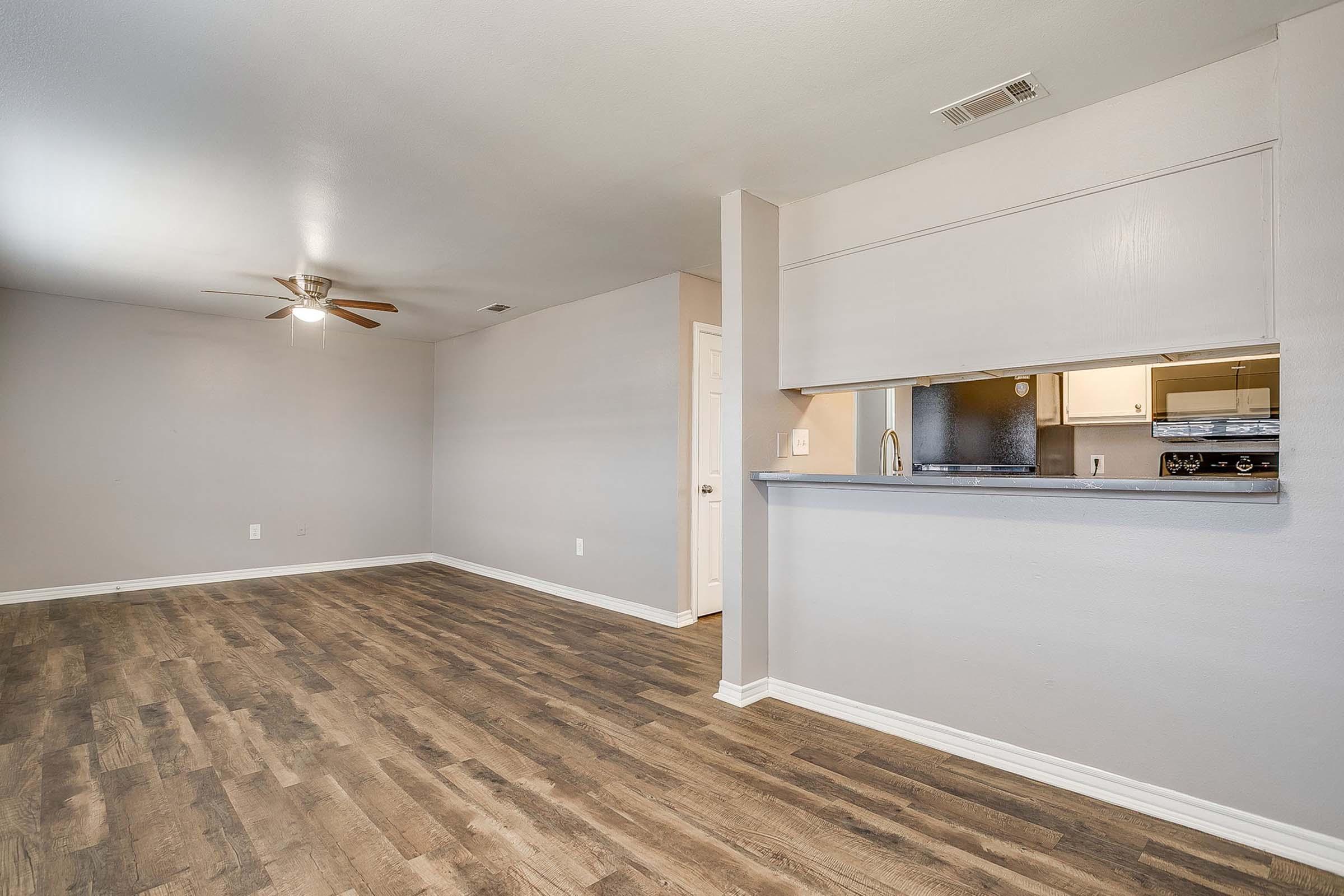 A spacious, empty living area with a light grey wall and wood-like flooring. A ceiling fan is mounted on the left side, and there's an open kitchen area visible through a pass-through counter. Natural light fills the room, creating a bright and inviting atmosphere.