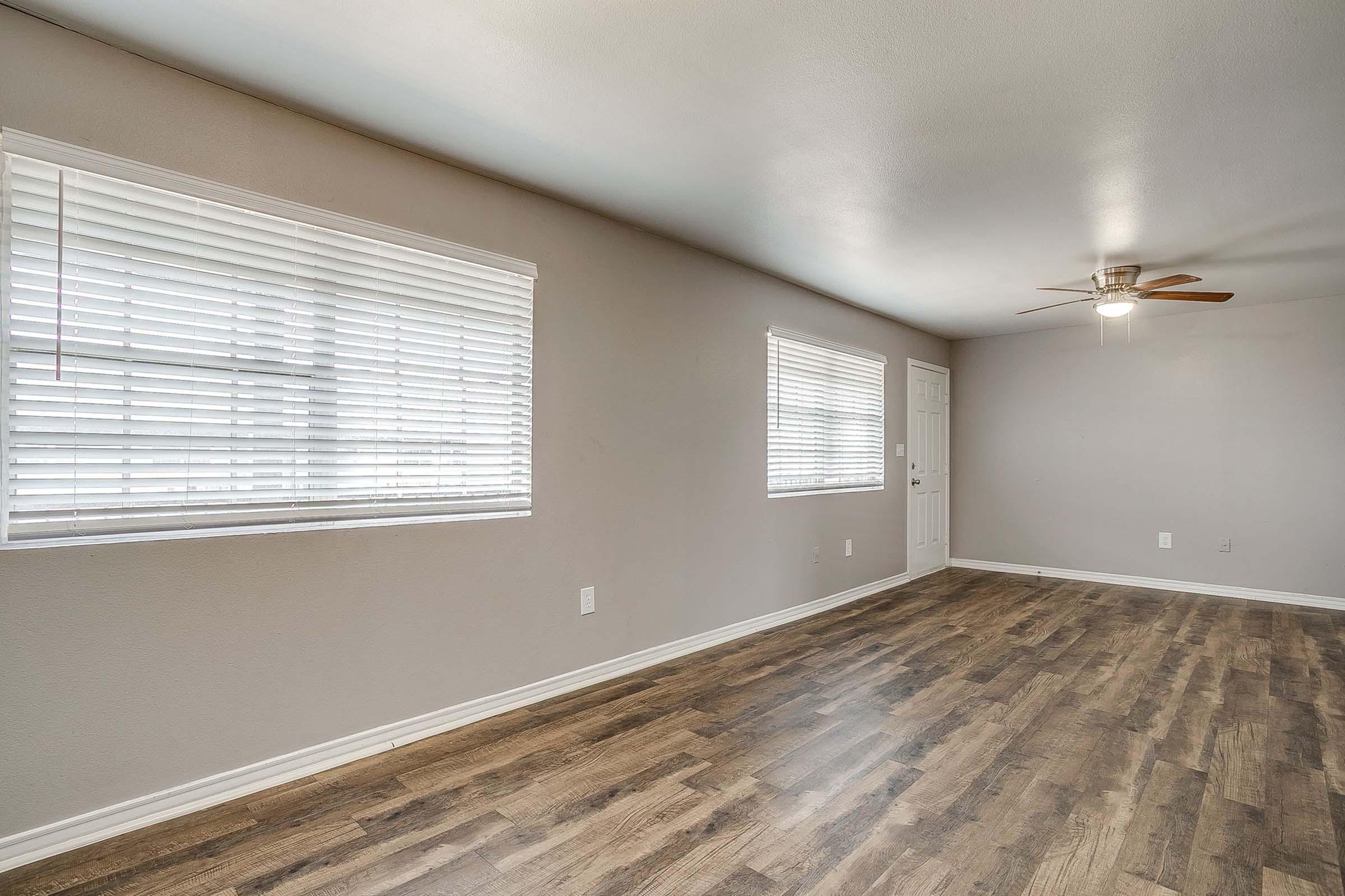 A spacious, empty room with light-colored walls and wooden flooring. Two windows with white blinds allow natural light to enter, and a ceiling fan is visible. A white door is situated on one side, creating a bright and airy atmosphere in the space.