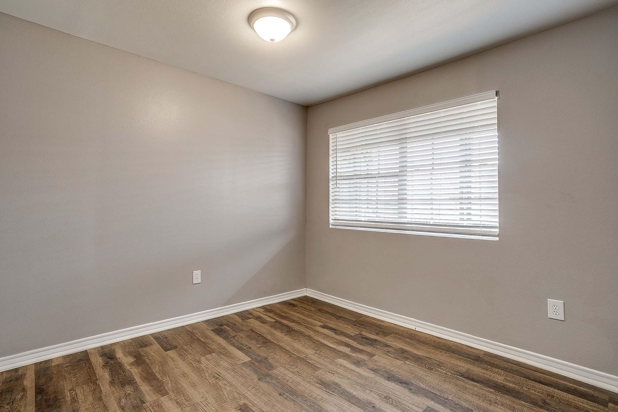 Empty room featuring light gray walls, wooden flooring, and a window with blinds. The ceiling is lit by a round light fixture, creating a neutral and spacious atmosphere.