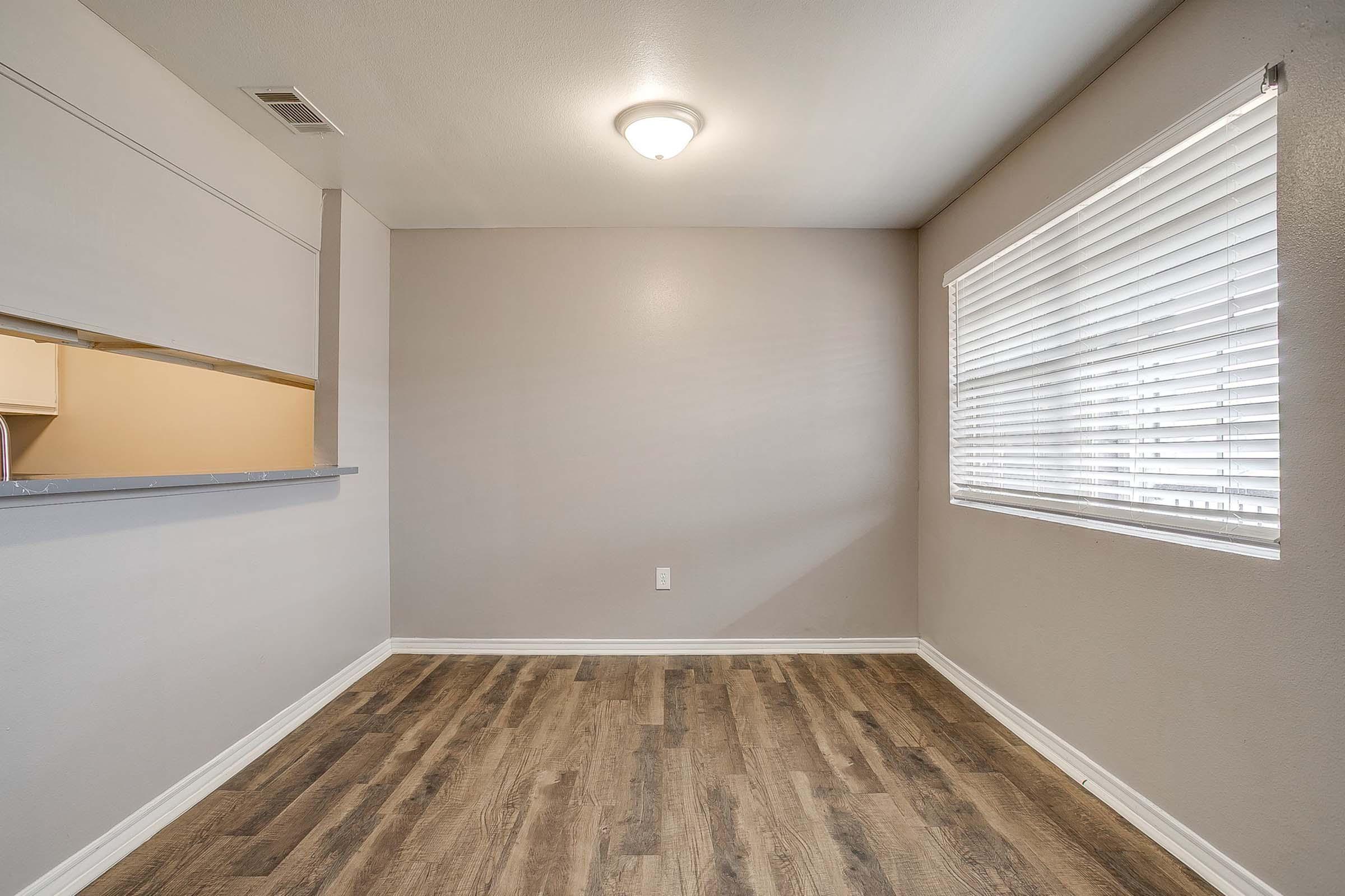 A small, empty room with light gray walls and a wooden floor. There is a window with blinds allowing natural light to enter, and a ceiling light fixture. A partial wall separates the room from a kitchen area, which is not visible. The space appears clean and ready for furniture.