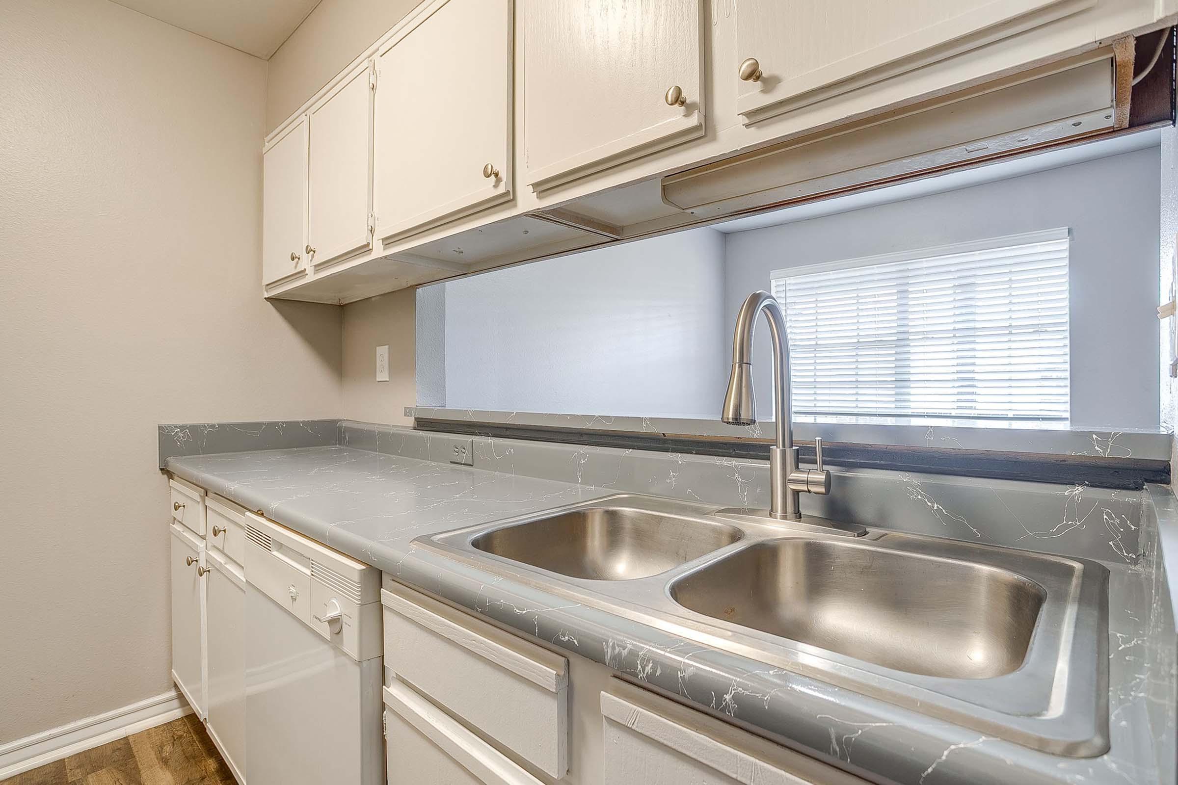 A kitchen with white cabinets, a gray stone countertop, and a double sink. The sink has a modern faucet, and there is a window with blinds above the sink, allowing natural light to brighten the space. The kitchen has a clean and functional appearance.