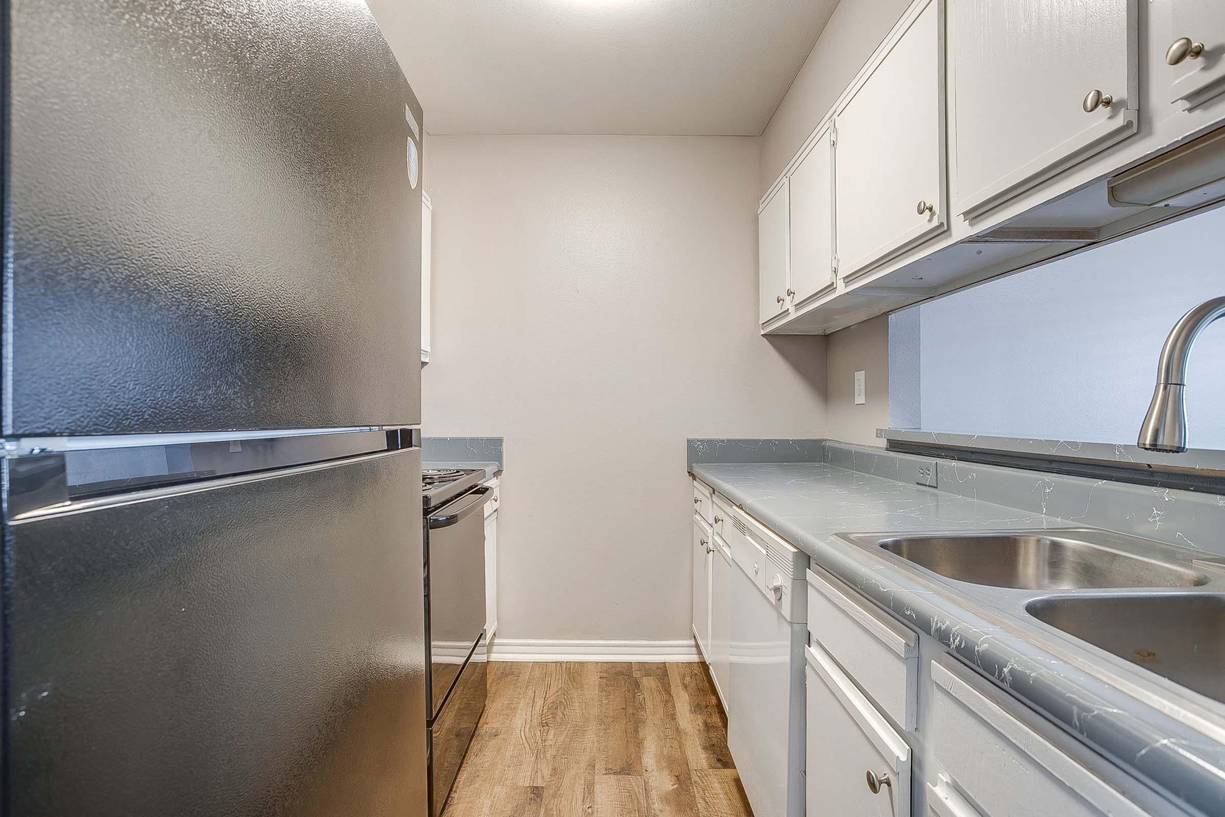 A modern kitchen featuring a black refrigerator, a white stove, and a sink with two basins. The cabinets are white with a light gray countertop. The flooring is a wood-like laminate. The kitchen is well-lit and has a clean, contemporary design.