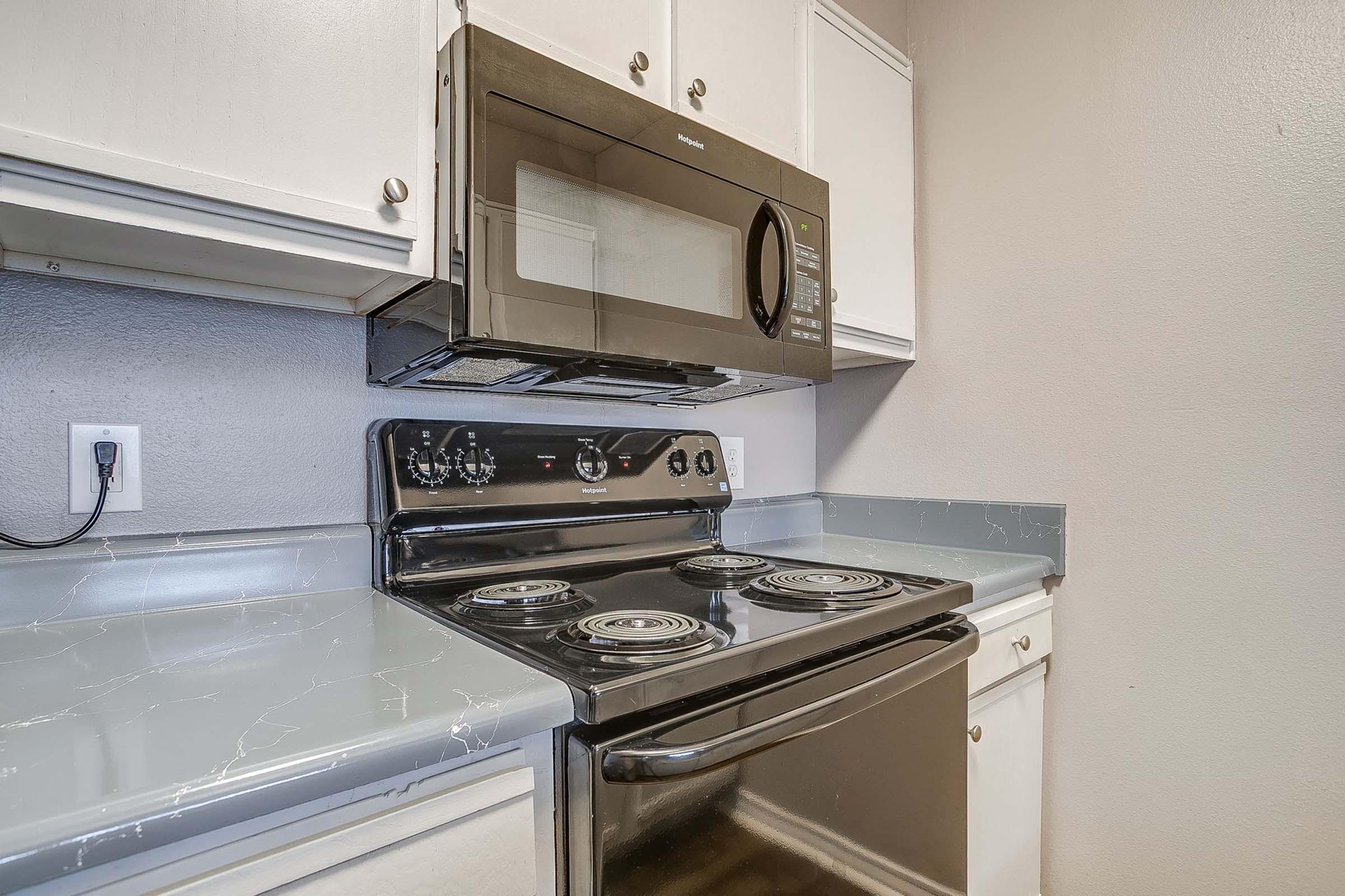 A modern kitchen featuring a black microwave mounted above a black electric stovetop. The stovetop has four burners and is situated on a gray countertop with a slight marble design. White cabinets are above the countertop, providing storage space. The kitchen has neutral-colored walls.
