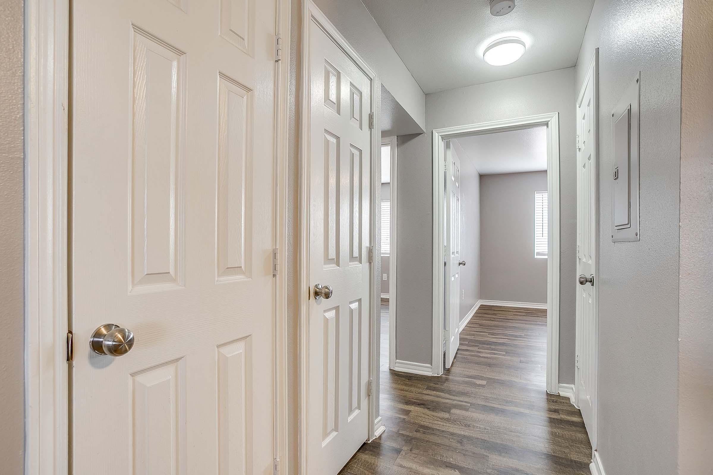 A view of a modern interior hallway featuring multiple white doors on either side. The walls are painted in a light gray color, and there are hardwood floors. Natural light filters in from a room at the end of the hallway, enhancing the spacious and inviting atmosphere.