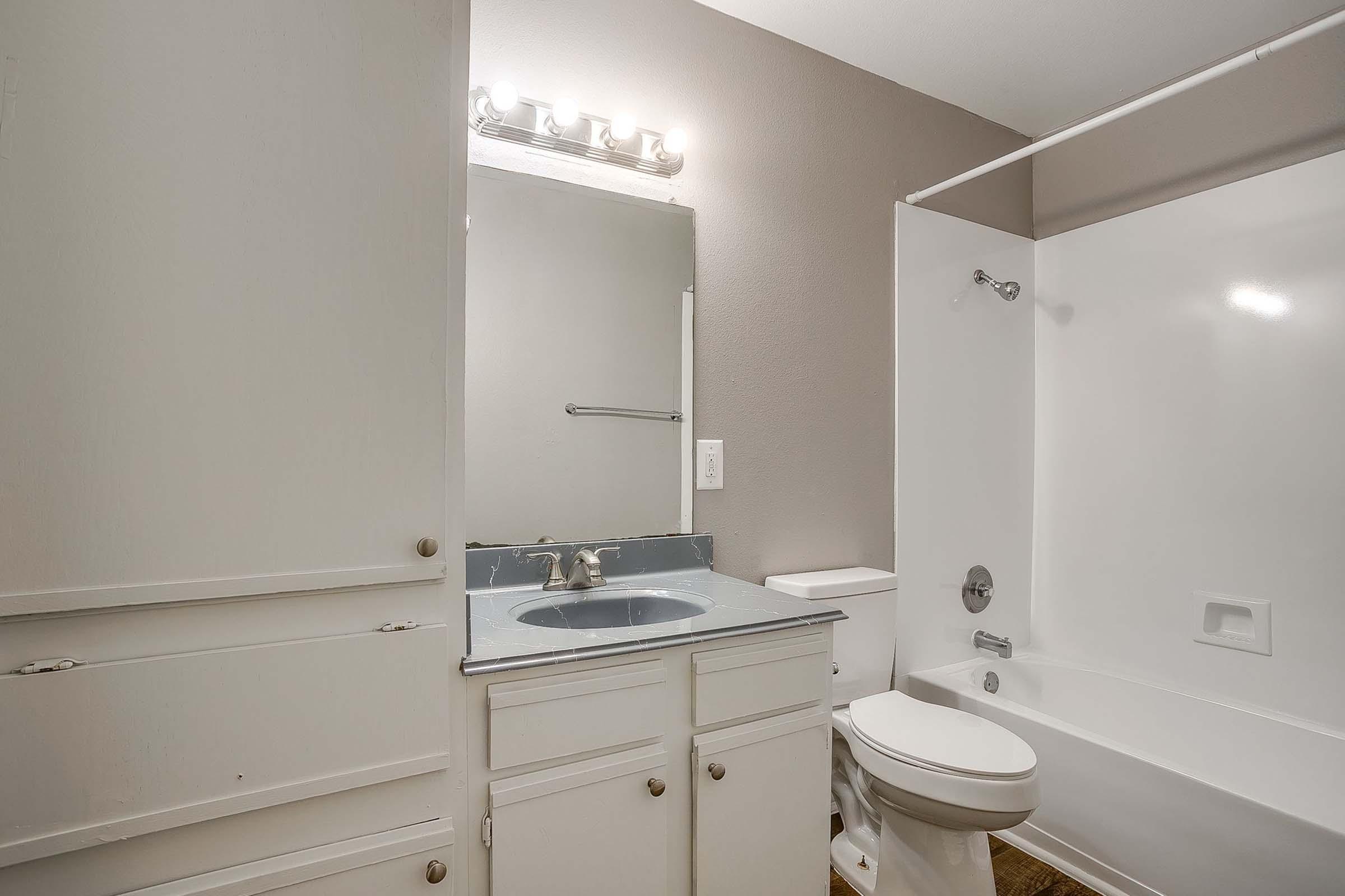 A clean and modern bathroom featuring a white tub and shower, a white toilet, and a sink with a blue countertop. The walls are painted a neutral gray, and there is a large mirror above the sink. The lighting consists of five bulbs above the mirror, providing bright illumination. Storage cabinets are visible on the left.