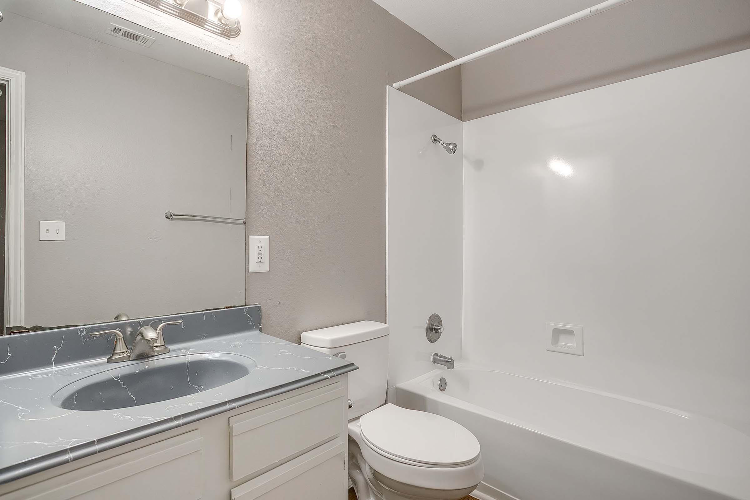 A clean and modern bathroom featuring a white bathtub and shower combo, a light gray countertop with a sink, a white toilet, and a large mirror above the sink. The walls are painted a soft gray, and there is good lighting from a fixture above the mirror.