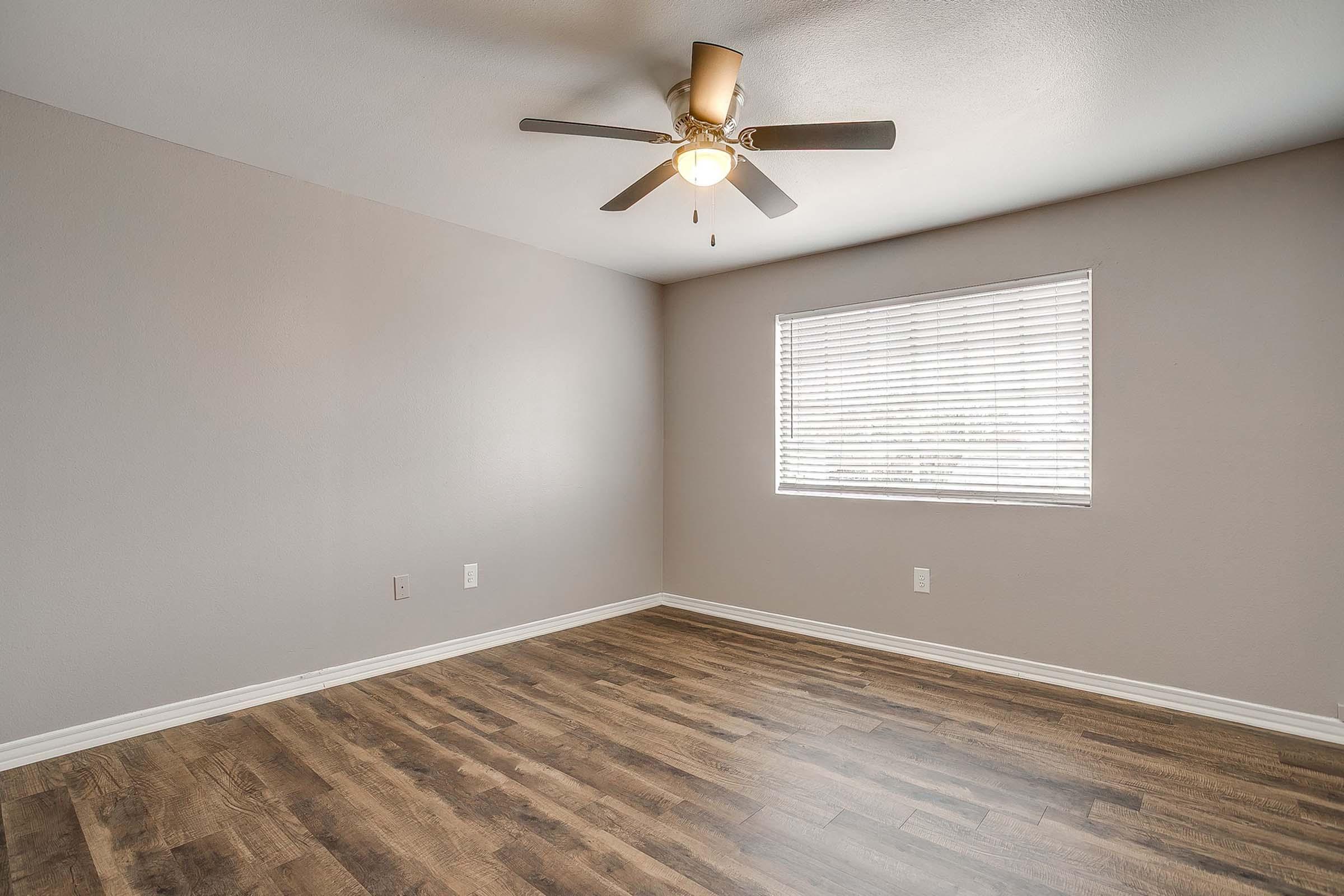 A vacant room with light gray walls and a ceiling fan. It features a window with blinds that lets in natural light, and has wood-like flooring. The room is empty, providing a spacious feel.