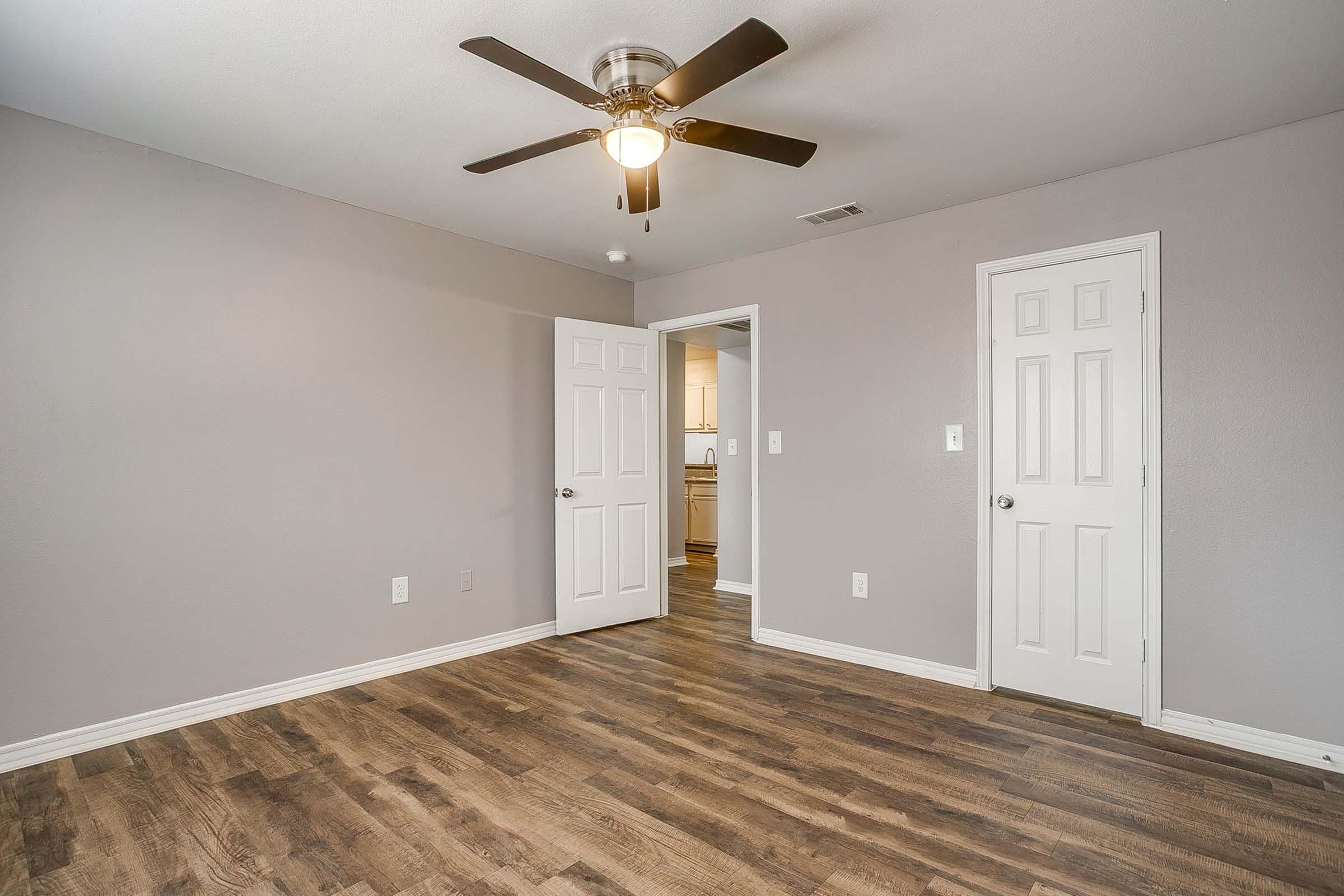 A well-lit, empty room featuring gray walls, a ceiling fan, and hardwood flooring. Two doors are visible, leading to other areas. The room has a clean, modern aesthetic with a spacious feel, perfect for potential furniture arrangement or decorating.