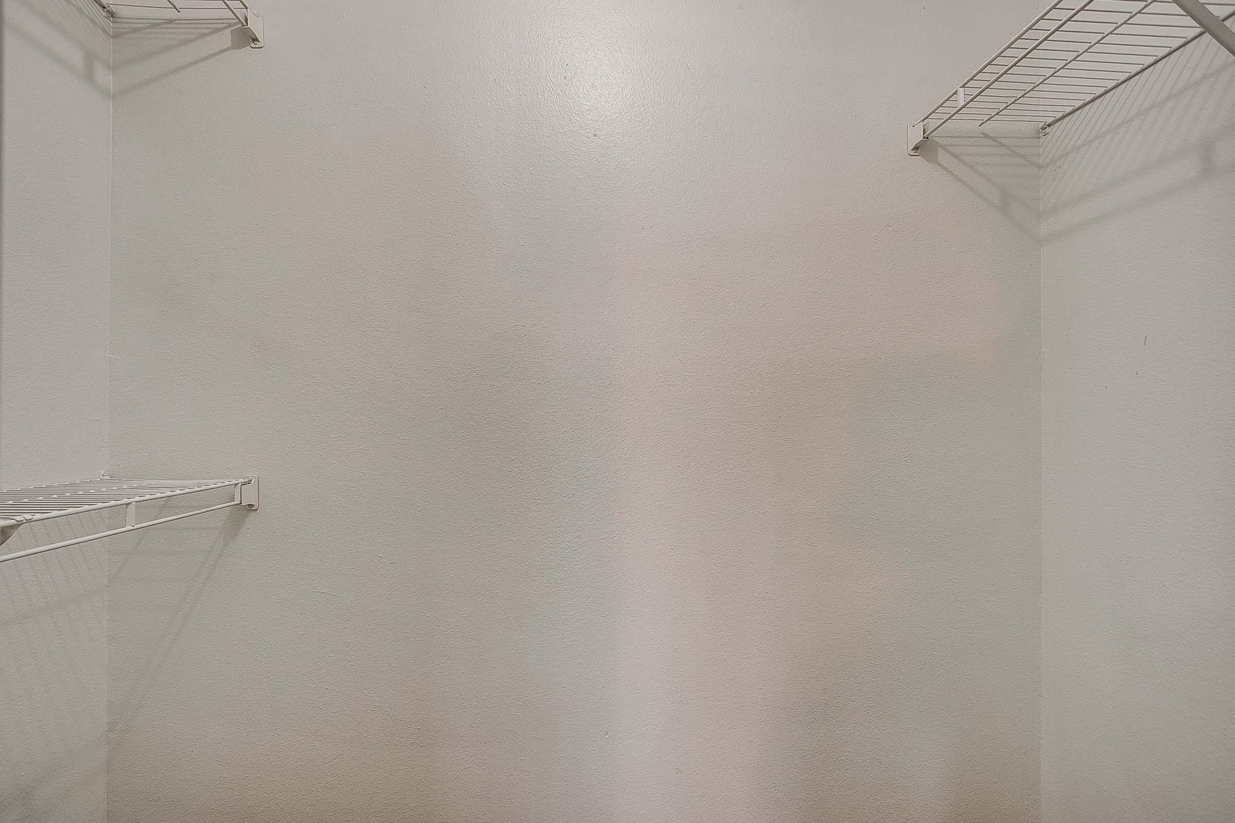 Empty closet with white walls and two wire shelves on either side. The shelves are suspended, and the walls have slight discoloration or stains. Overall, the space appears unoccupied and minimalistic.