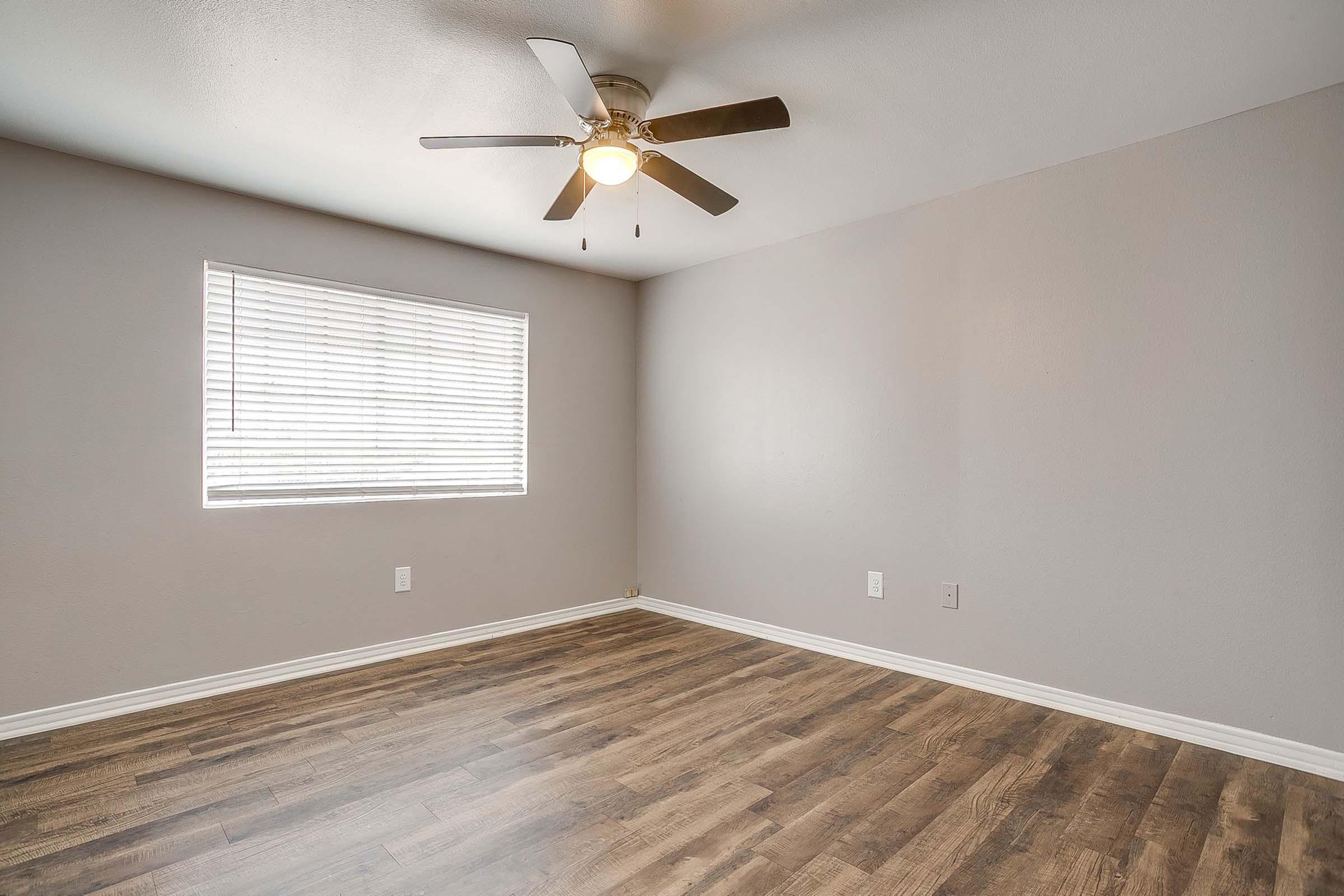 Empty room with light gray walls and wooden flooring. A ceiling fan with a light fixture is mounted in the center. One window with white blinds lets in natural light, casting soft shadows on the floor. The space is minimalistic and modern, with no furniture or decoration.