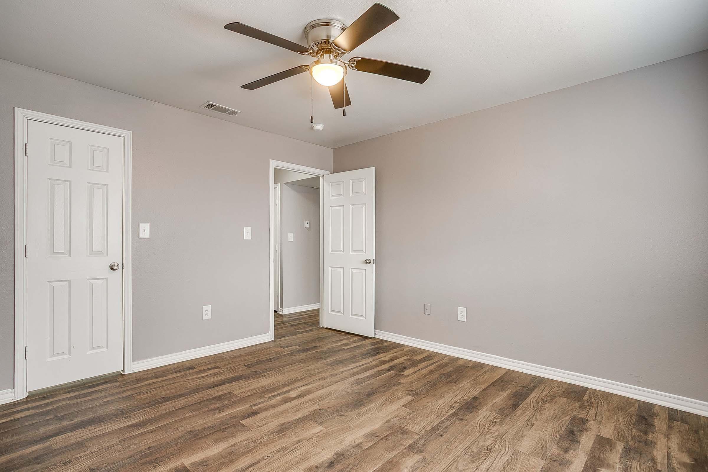 A spacious, minimally decorated room with gray walls and a ceiling fan. There are two doors, one leading to another room and the other to a closet. The floor is made of wood laminate, enhancing the modern look of the space. Natural light is softly coming in, creating a bright atmosphere.