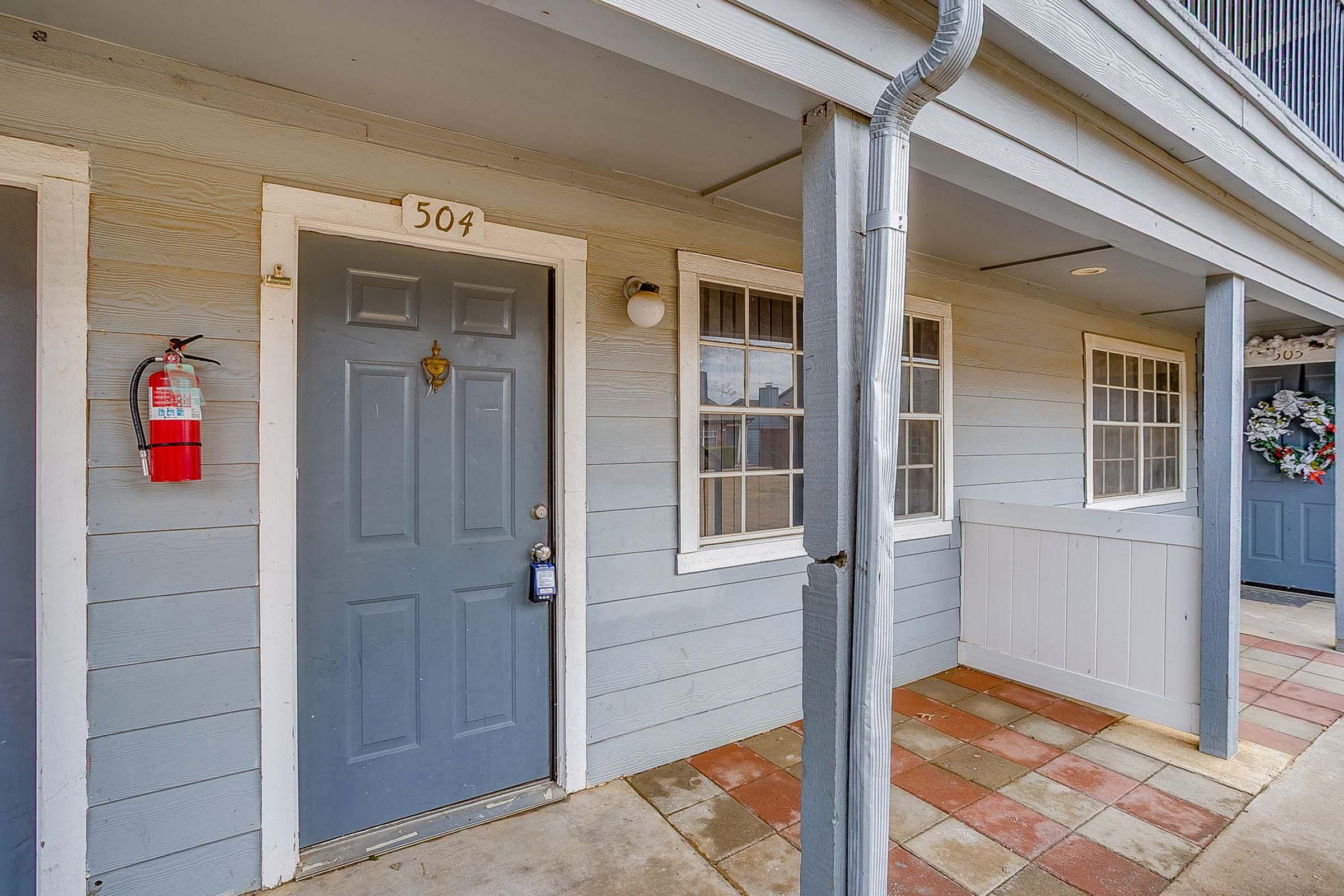 A gray door labeled "504" with a brass door handle, located in a corridor of a low-rise building. Two windows beside the door and a fire extinguisher mounted on the wall. The entrance features a covered area with tiled flooring, and a decorative wreath hangs on the opposite side.