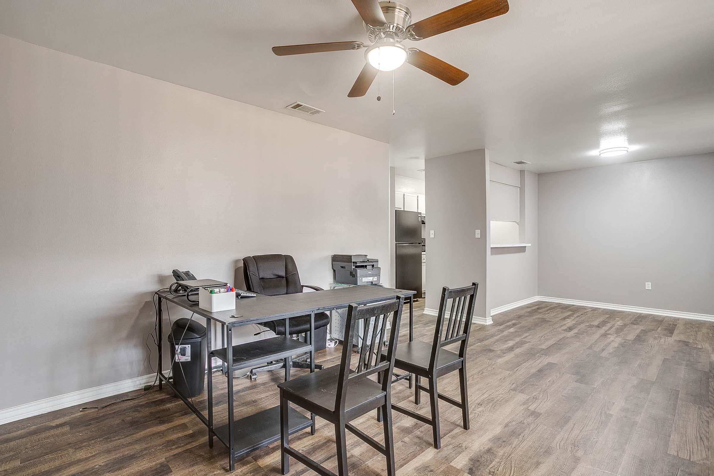 A simple living or office space featuring a ceiling fan, a wooden floor, a desk with a phone and office equipment, and two black chairs. The walls are painted light gray, and there is a kitchen area visible in the background. Natural light brightens the room.
