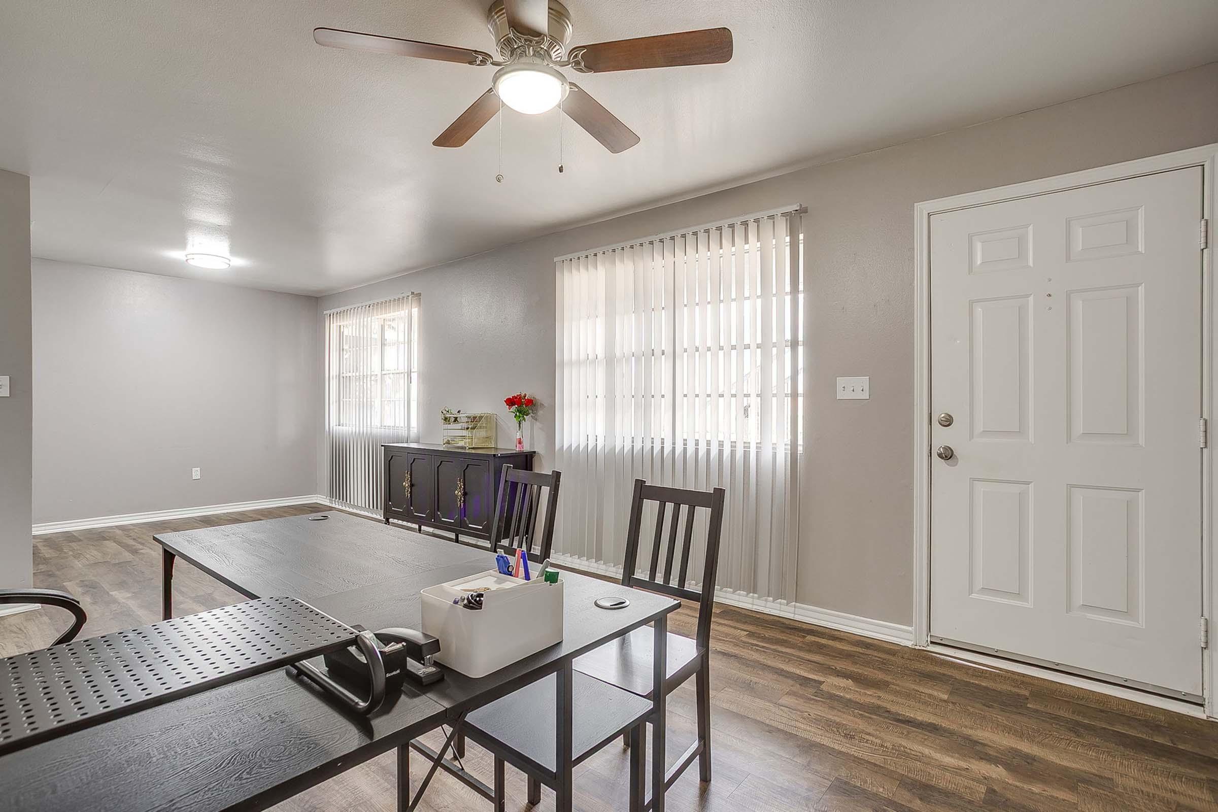 A modern living space featuring a dark table with chairs, a ceiling fan, and large windows with blinds. An entry door is visible, along with a small table holding a phone and office supplies. The walls are painted in a light color, creating a bright and airy atmosphere.
