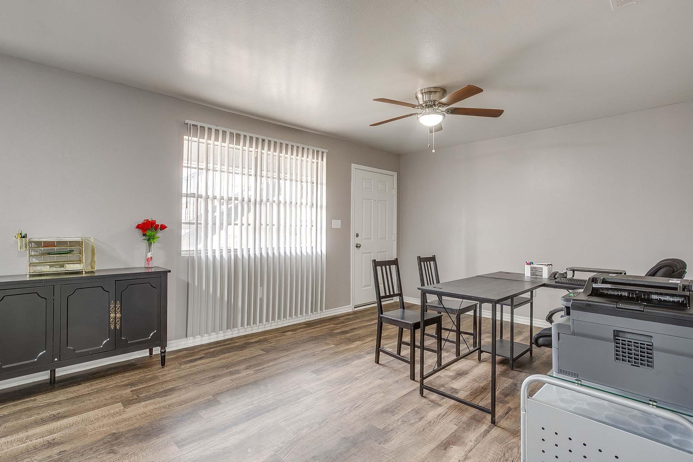 A bright and spacious room with gray walls and a wood-like floor. A table with four black chairs is in the center, alongside a black cabinet and a printer. A door leads outside, while a window with vertical blinds allows natural light in. A small vase with red flowers adds a pop of color.