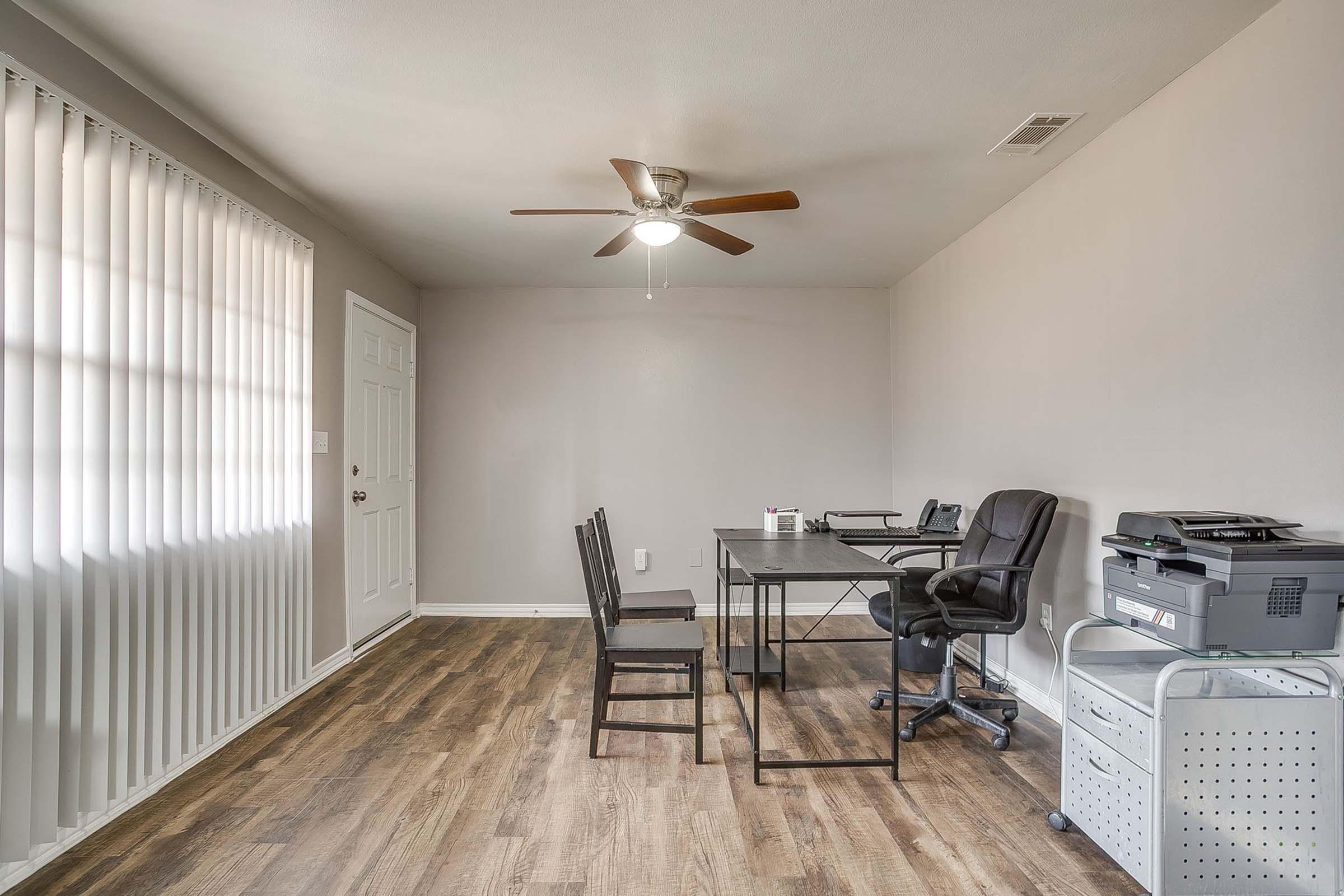 A simple, well-lit home office featuring a wooden floor, a ceiling fan, and a desk with two chairs. There is a printer on a cart and a closed door visible. The walls are painted a light color, and large windows have vertical blinds, allowing natural light to enter the space.