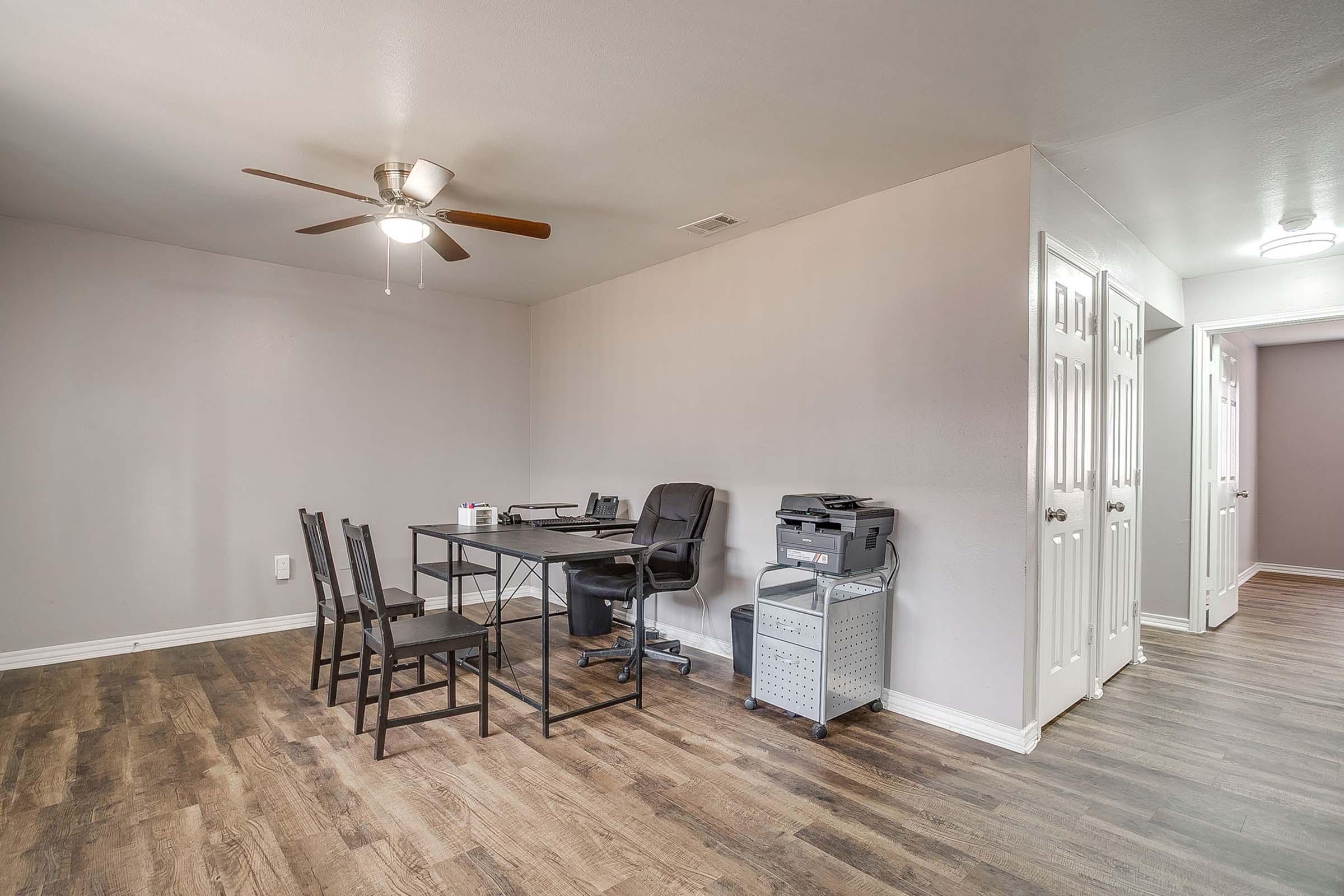A minimalist interior room with light gray walls and wooden flooring. There is a small office setup featuring a table, black office chair, and a printer. Nearby are four black chairs and a ceiling fan. Two closed doors can be seen in the background, adding a functional touch to the space.