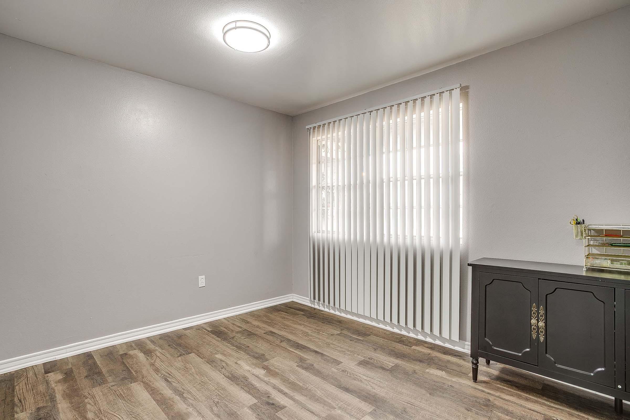 Empty room with light gray walls and wooden flooring. A window with vertical blinds allows natural light to enter. A modern ceiling light is installed. A black furniture piece with decorative hardware is against one wall, adding a touch of elegance to the minimalist space.