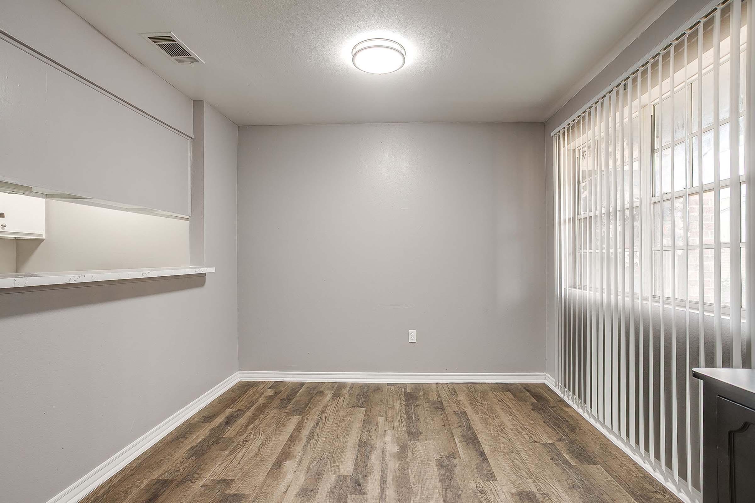 A small, empty room featuring light gray walls and a hardwood floor. There is a window with vertical blinds allowing natural light and a ceiling light fixture. A partial wall separates the room from another space, suggesting it may be part of a larger area, like a living room or kitchen.