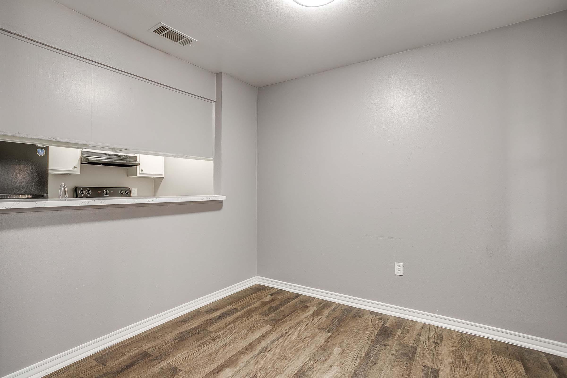 A spacious, empty room with light gray walls and a hardwood floor. There is a small pass-through kitchen area visible with white cabinetry and kitchen appliances. The room is well-lit, with a ceiling light fixture, and has no furniture, highlighting its open space.