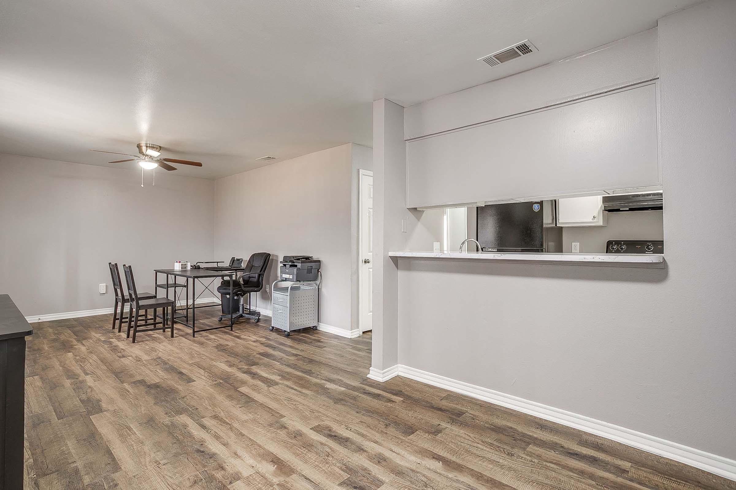 A spacious living area featuring hardwood-style flooring, a ceiling fan, and a kitchenette visible through an open bar. There is a dining table with four chairs, a black desk with a swivel chair, and a printer, creating a functional workspace. Neutral walls contribute to a bright, airy atmosphere.