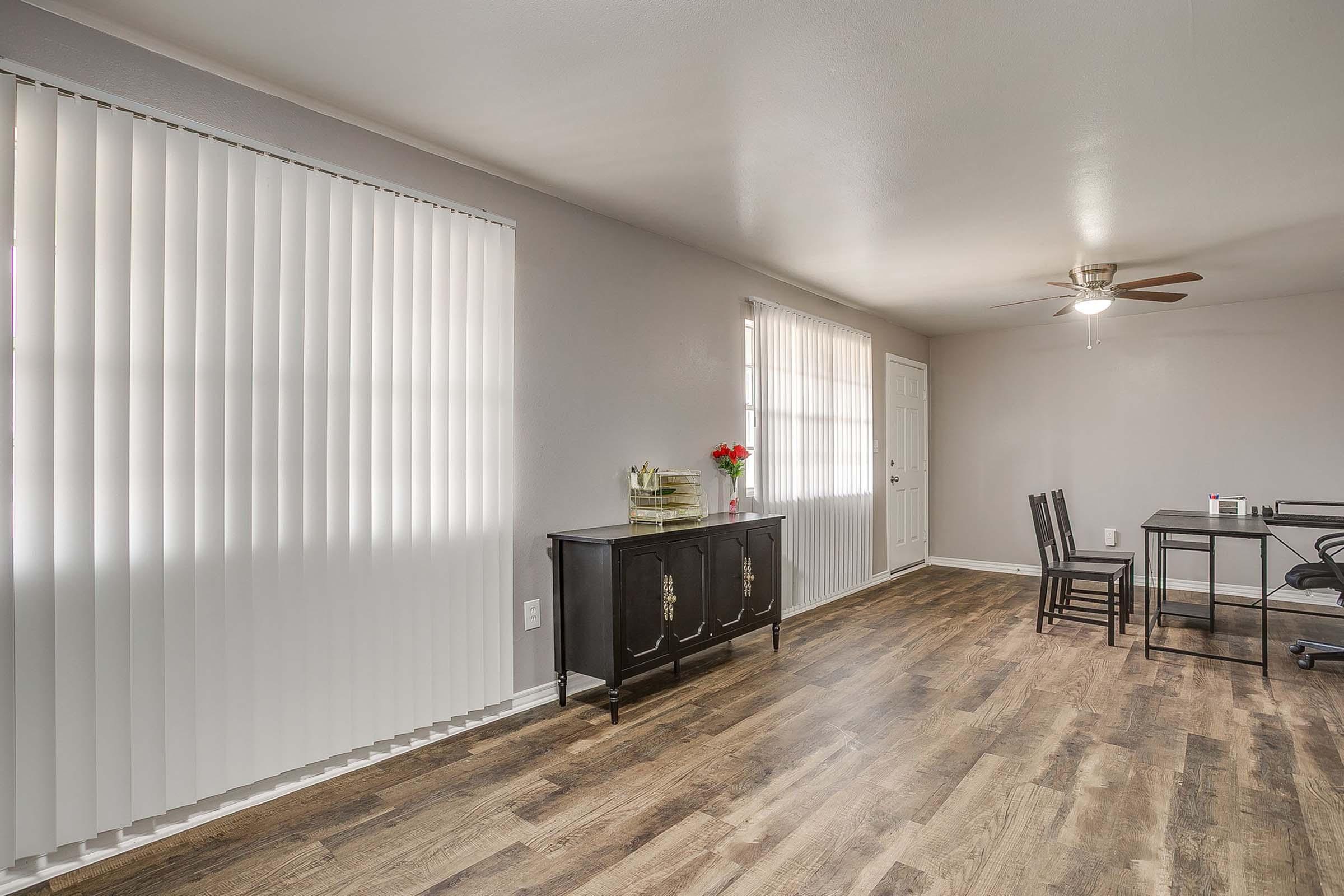 A spacious, well-lit living area featuring light-colored walls, vertical blinds on large windows, a dark wooden sideboard, a small dining table with four chairs, and a ceiling fan. The floor has a warm-toned wood finish, and a door leads to the outside.