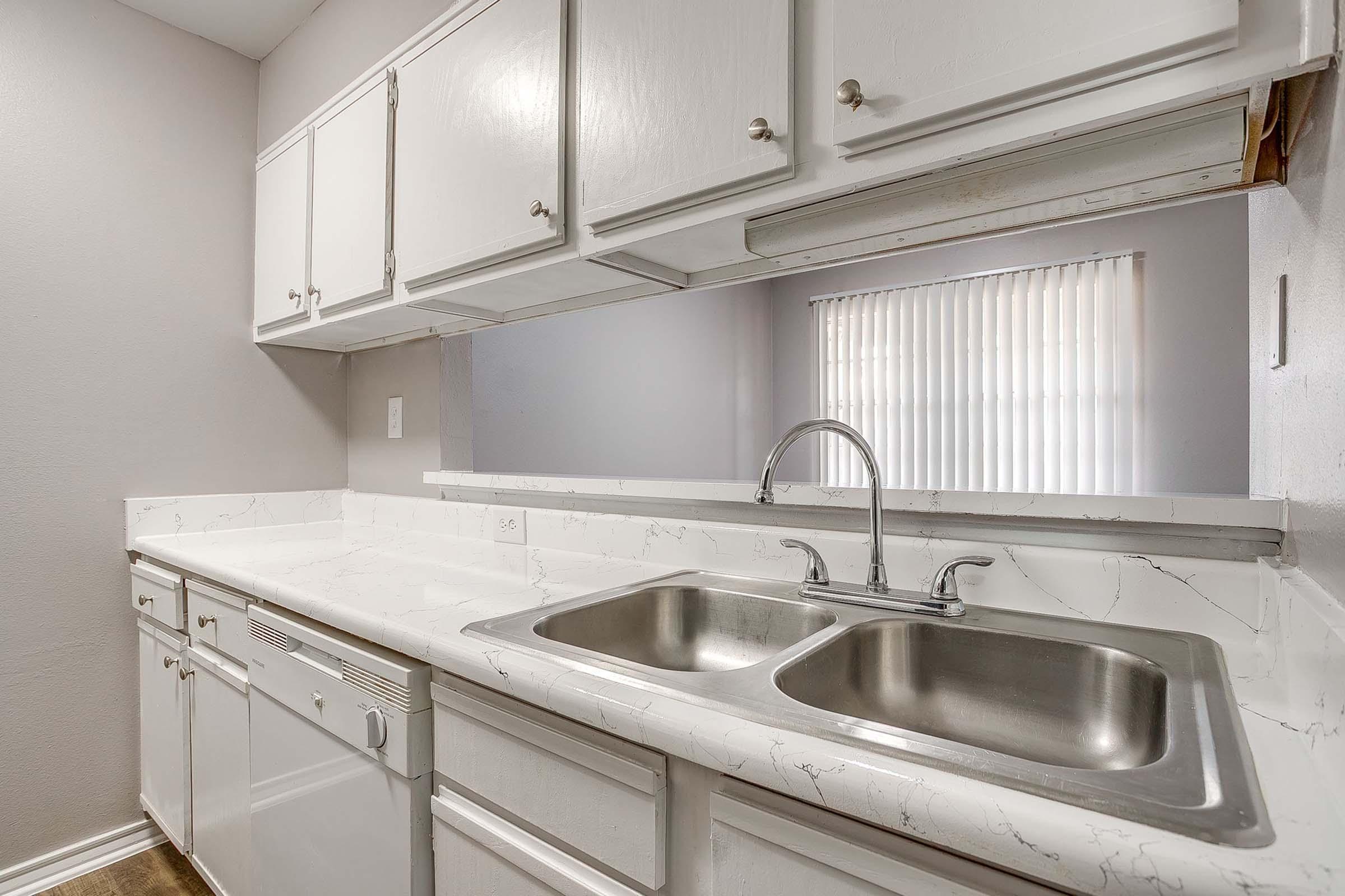 A modern kitchen featuring white cabinets, a double sink, and a marble-patterned countertop. The design is minimalist with a light gray wall in the background and a window covered by blinds providing natural light.