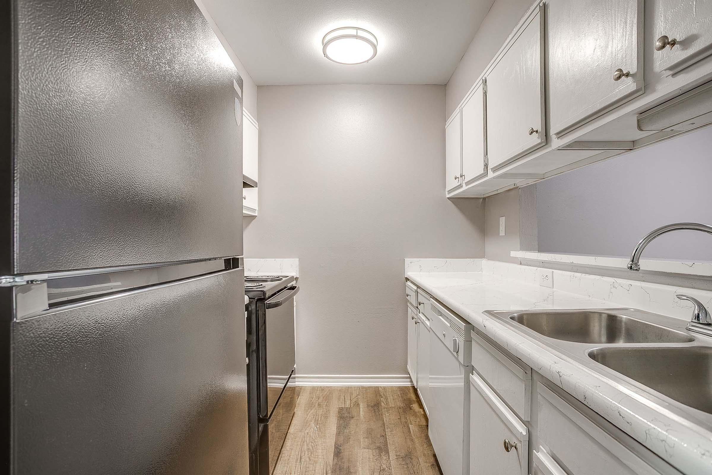 A small kitchen featuring stainless steel appliances, including a refrigerator and stove, white cabinetry, and a double sink. The walls are painted in a light color, and the floor is covered with wood-like laminate. A ceiling light fixture provides overhead illumination.