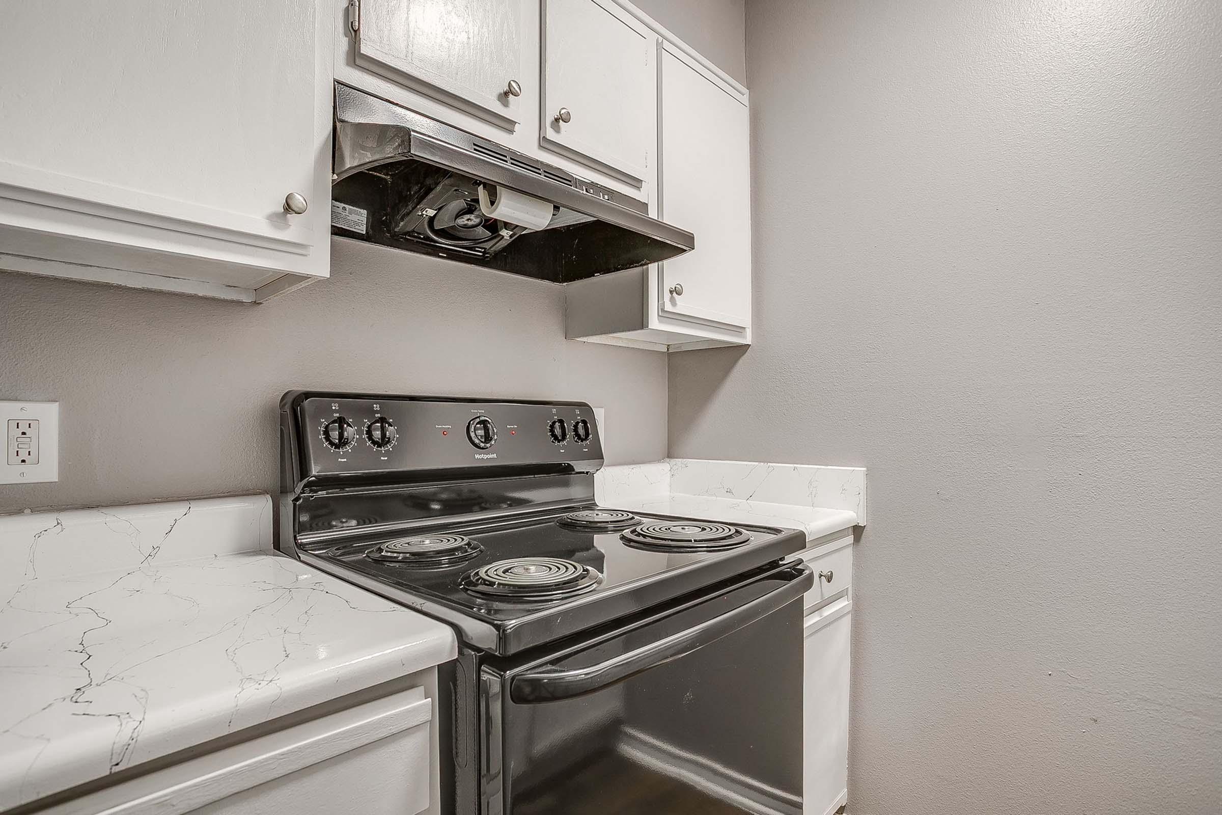 A compact kitchen featuring a black stove with four burners, an overhead vent hood, and white cabinets. The countertops have a marble-like finish, and the walls are painted in a neutral gray color, creating a modern and clean look.