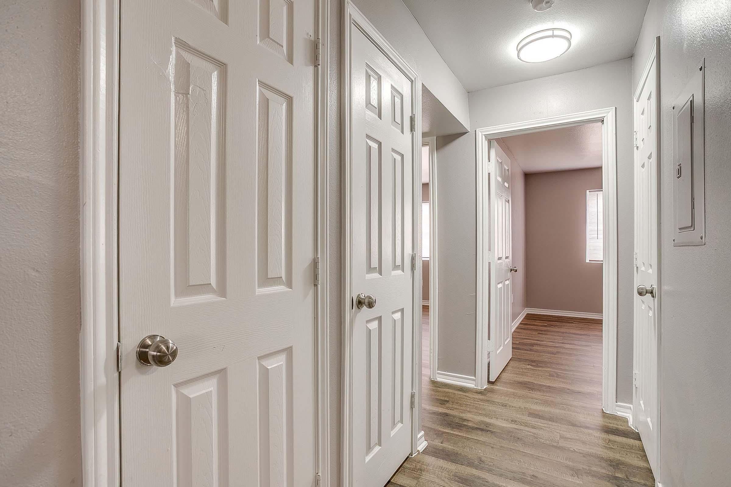 A well-lit hallway with light gray walls and wooden flooring. Two closed white doors are visible on the left, and an open doorway leads to another room on the right. Natural light filters in from a window at the end of the hall, creating a welcoming atmosphere. A ceiling light fixture is mounted above.