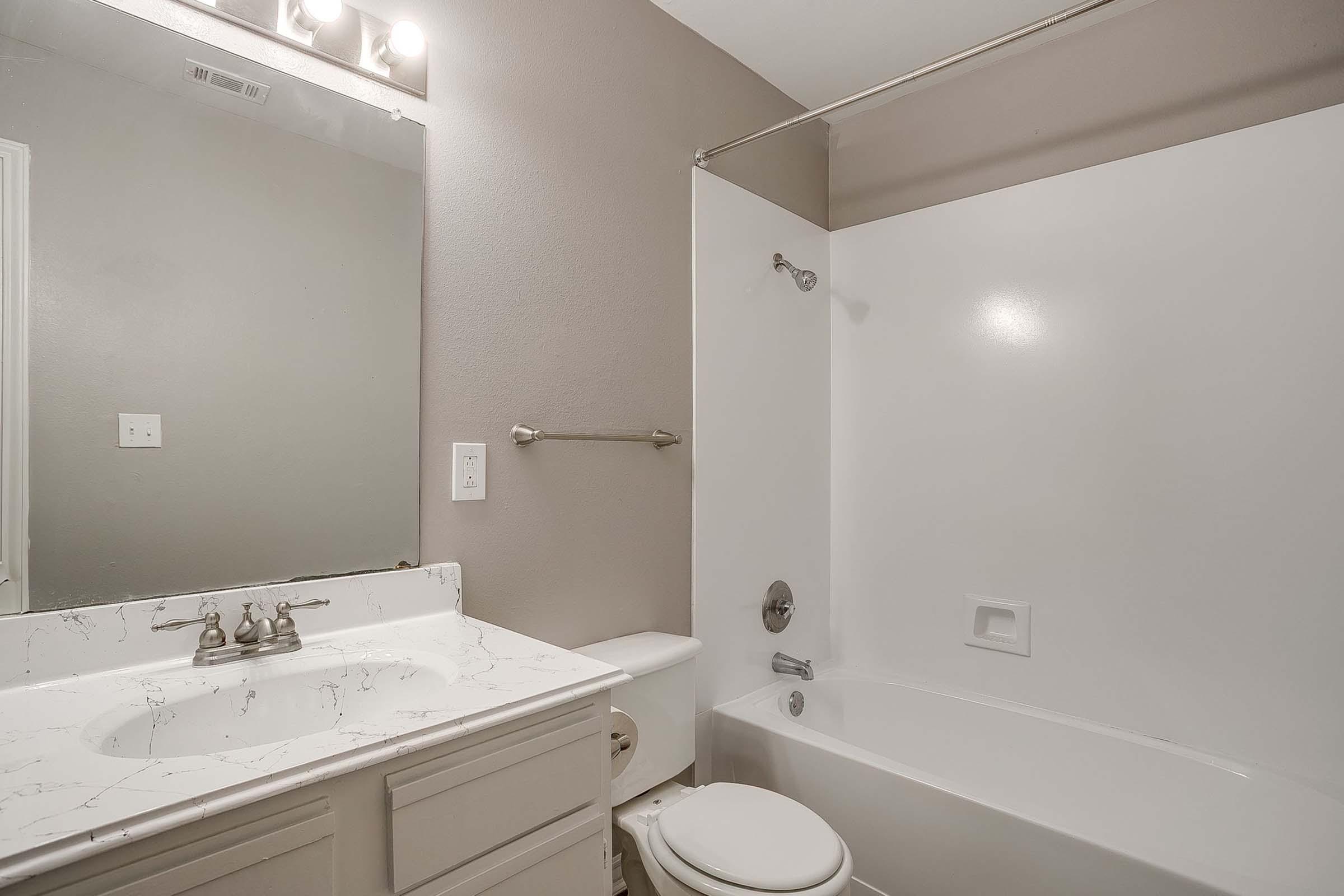 A clean, modern bathroom featuring a white bathtub and shower combination, a light-colored countertop with a sink, a large mirror, and neutral-colored walls. The space is well-lit with overhead fixtures, creating a fresh and inviting atmosphere.