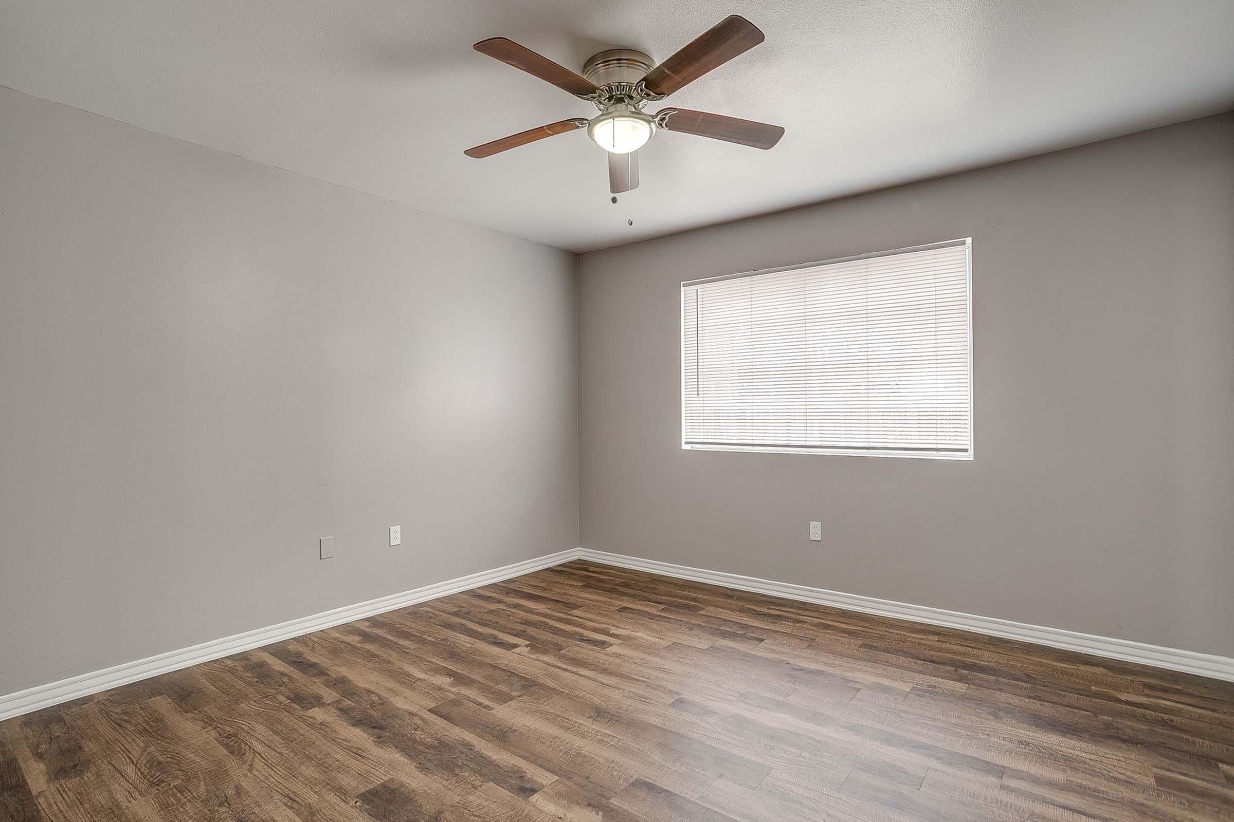 Empty room featuring light gray walls, a ceiling fan with wooden blades, and a window with blinds. The flooring is a warm-toned wood laminate, and there are no furniture or decorations in the space, creating a minimalist look.