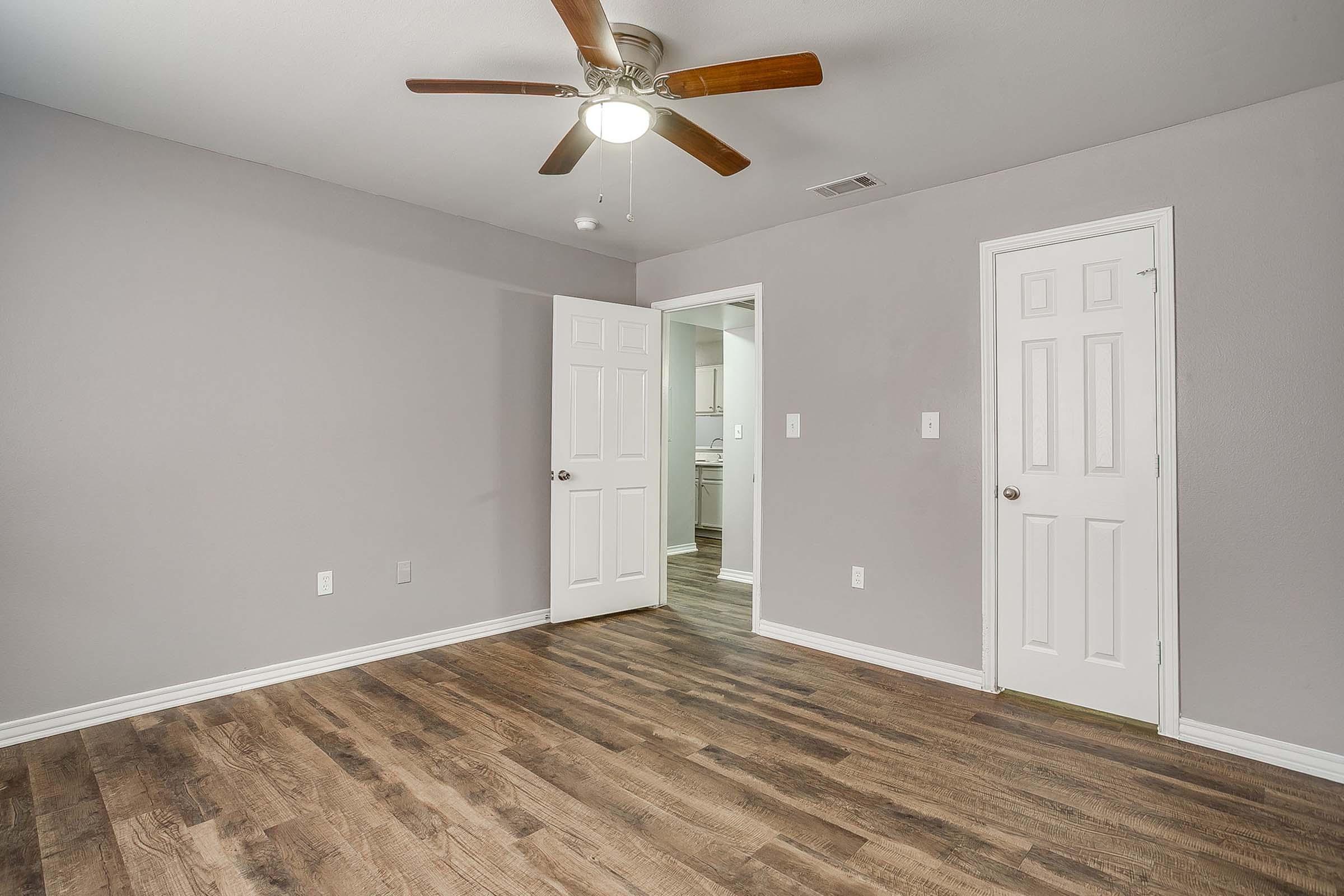 Interior view of a vacant room featuring a ceiling fan, gray walls, and hardwood-style flooring. Two white doors lead to different areas, with one door partially open, revealing a glimpse of a kitchen area beyond. The space is well-lit and clean, ideal for new occupants to personalize.