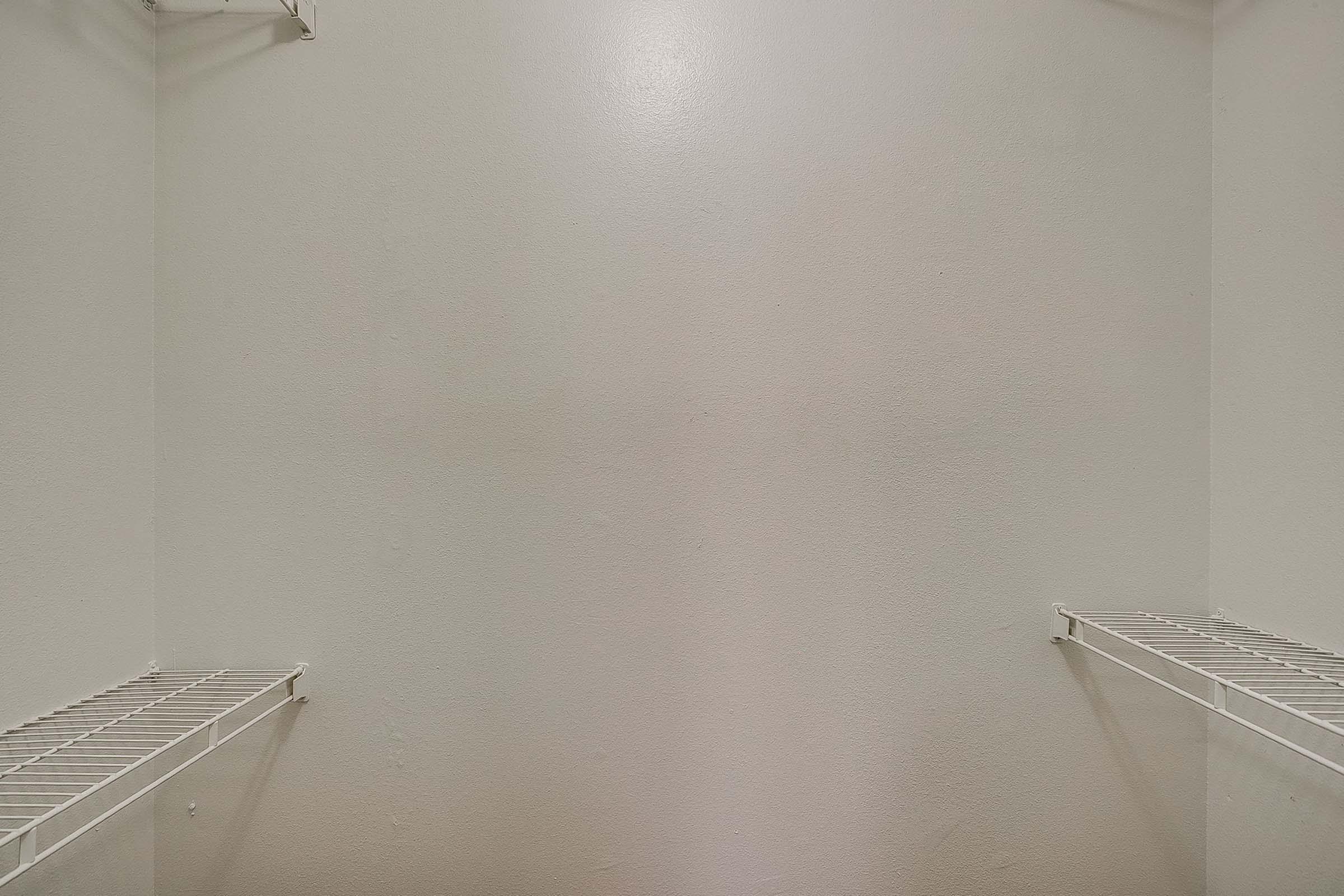 Empty closet space with two wire shelves on either side, featuring plain white walls. The overall atmosphere is clean and minimalistic, with no items stored inside.