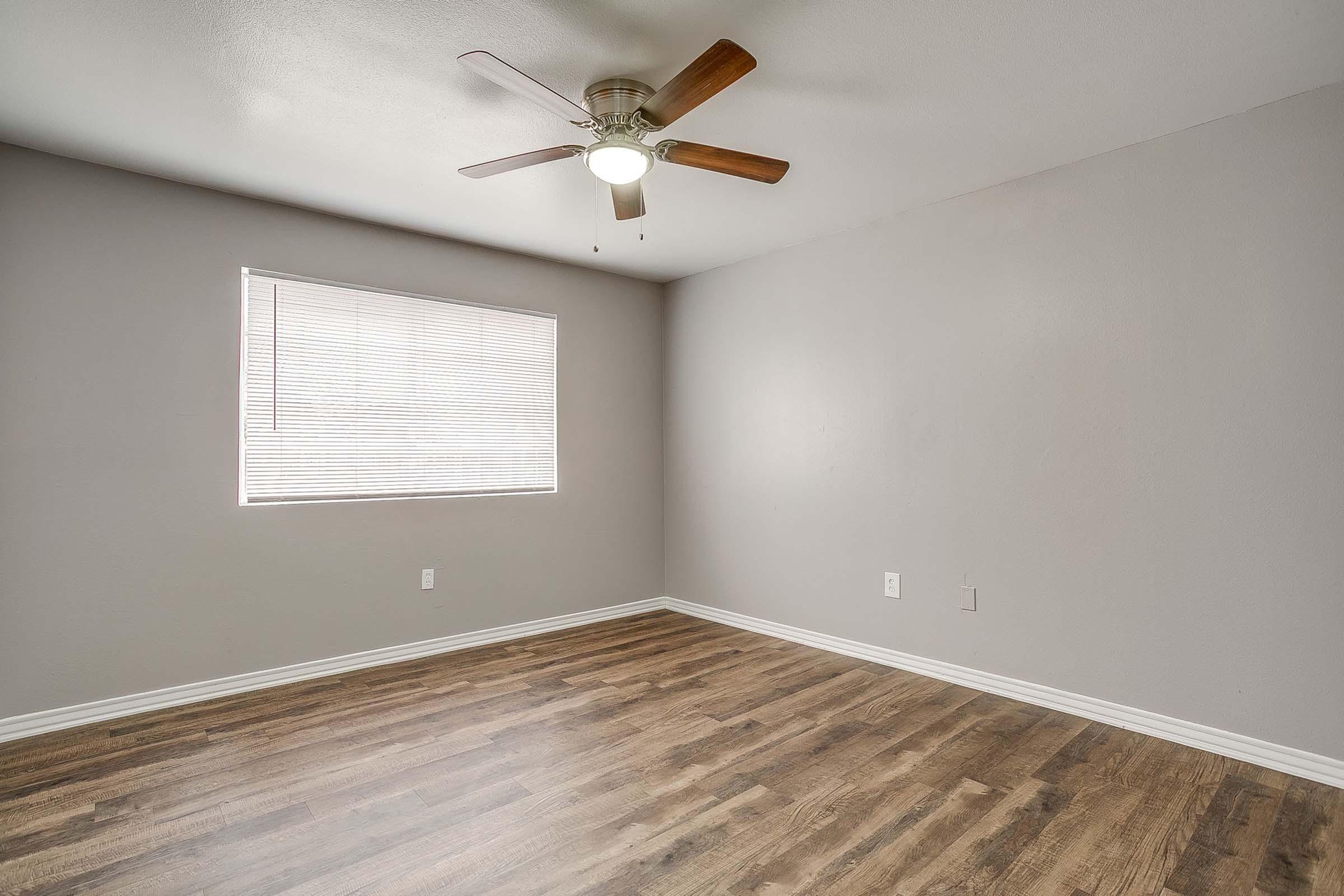 A bare room with light gray walls and wooden flooring. It features a ceiling fan with wooden blades and a window with white blinds allowing natural light to enter. The space is uncluttered and spacious, ideal for personal decoration or use as a living area.