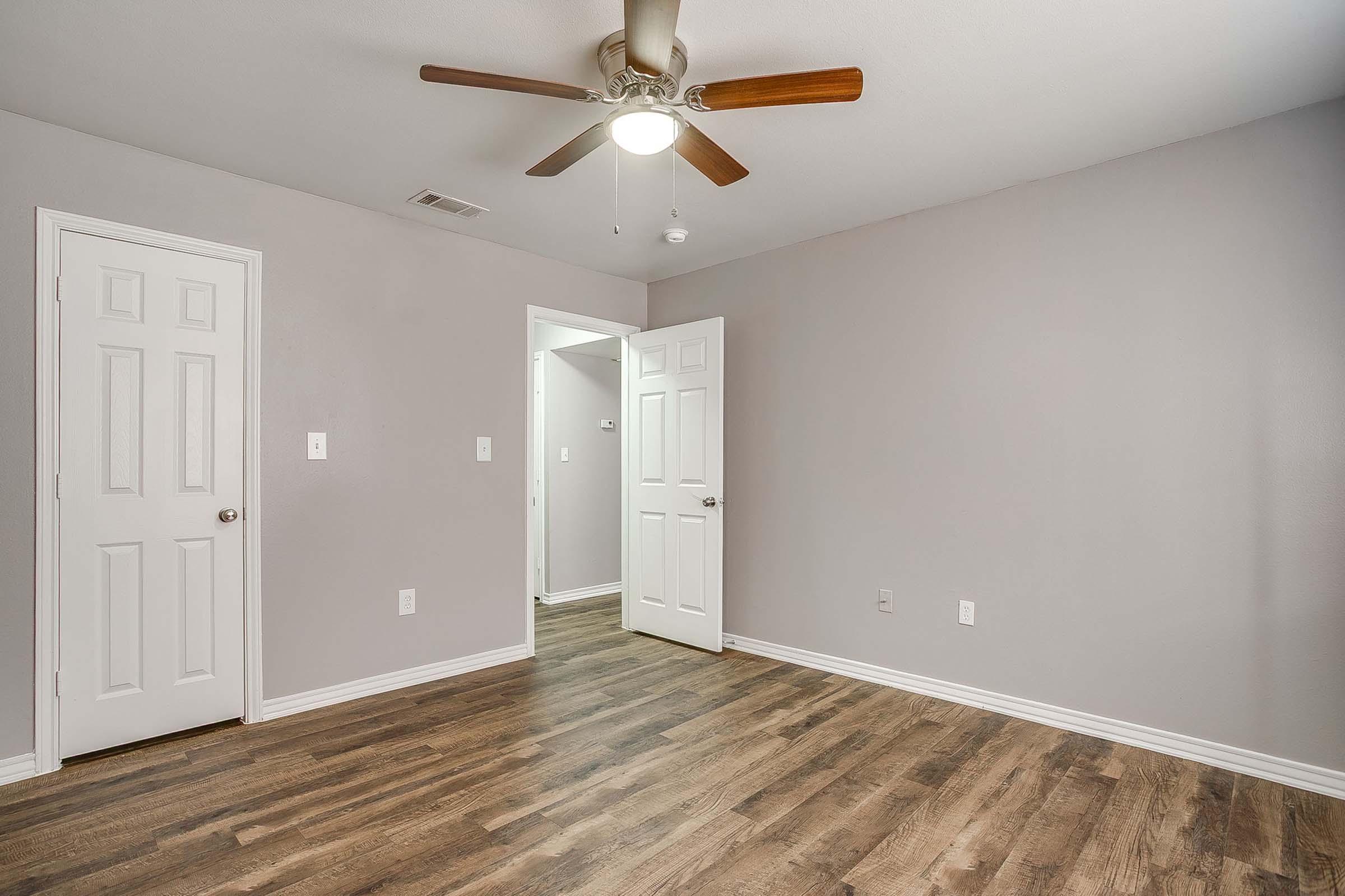 A spacious room with light gray walls, wooden flooring, and a ceiling fan. Two doors are visible, one leading to another room and the other to a closet. The room is brightly lit, creating a welcoming atmosphere.