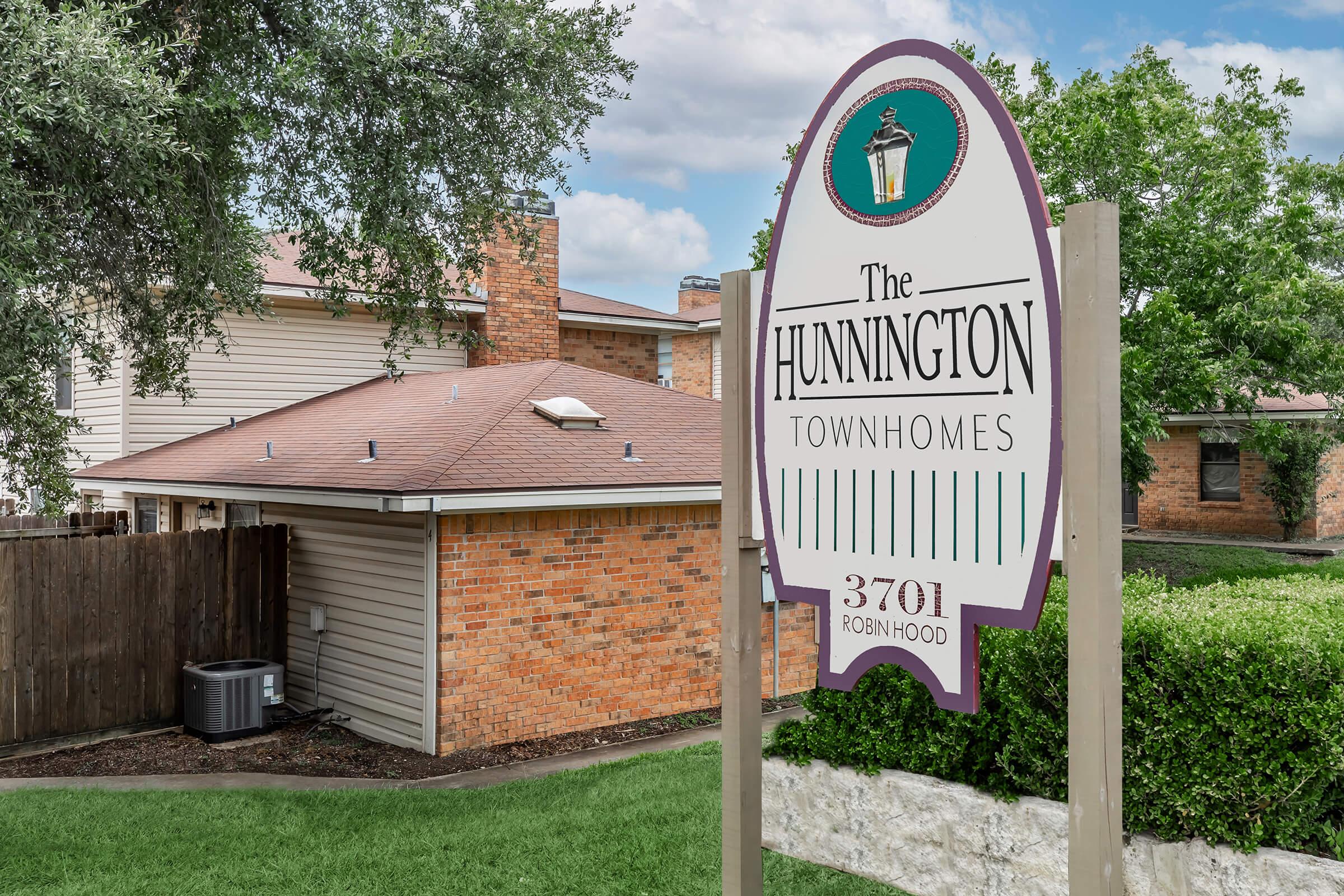 Sign for The Hunington Townhomes, featuring the name prominently at the top with a decorative lantern icon. The sign includes the address "3701 Robin Hood" below. Surrounding environment includes grassy areas and nearby residential buildings.