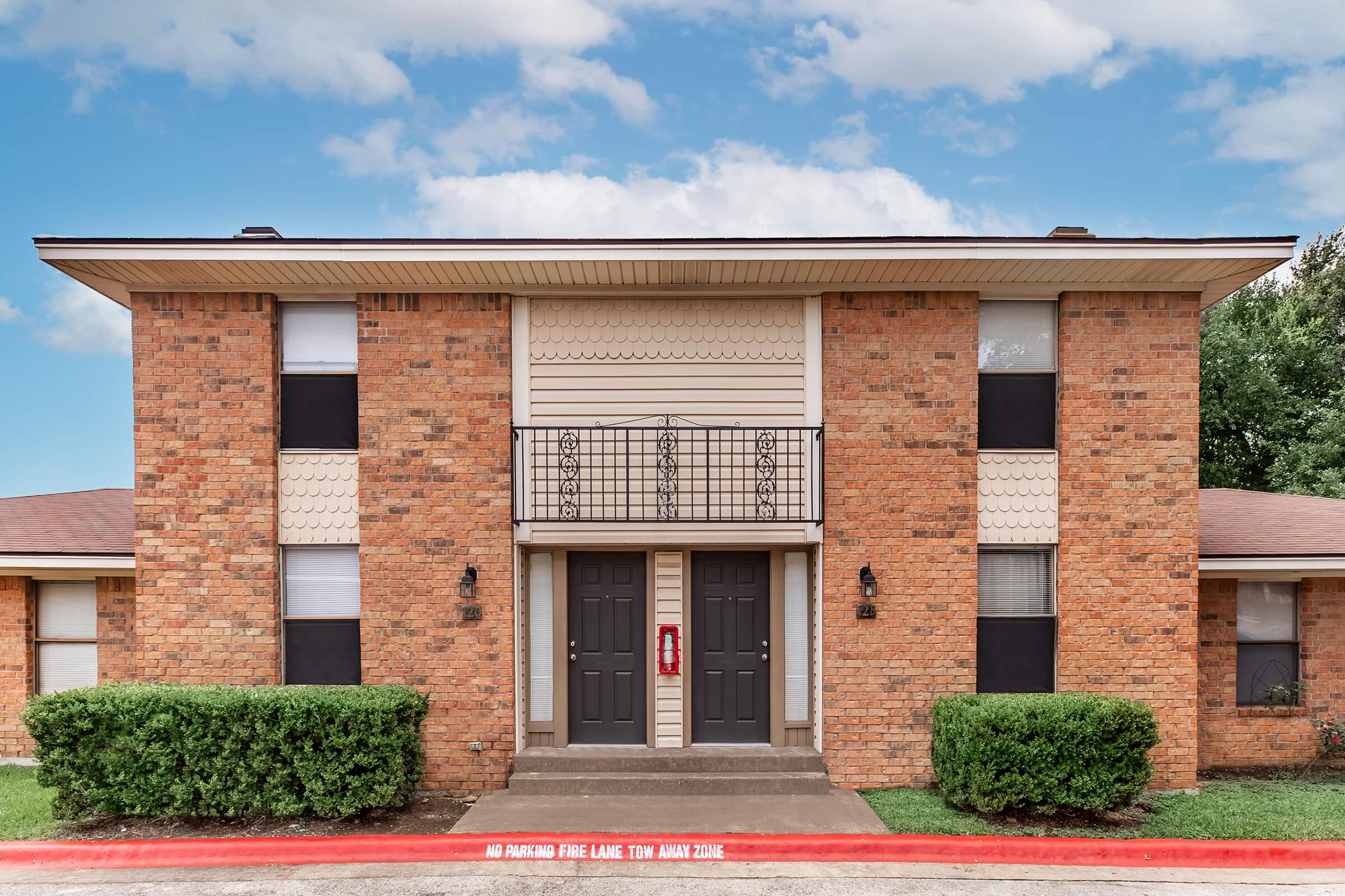 A two-story brick apartment building with a symmetrical design, featuring two front doors and a small balcony on the second floor. Lush green shrubs are planted in front, and there is a red pavement marking stating "No Parking Fire Lane Tow Away Zone." The sky is partly cloudy.