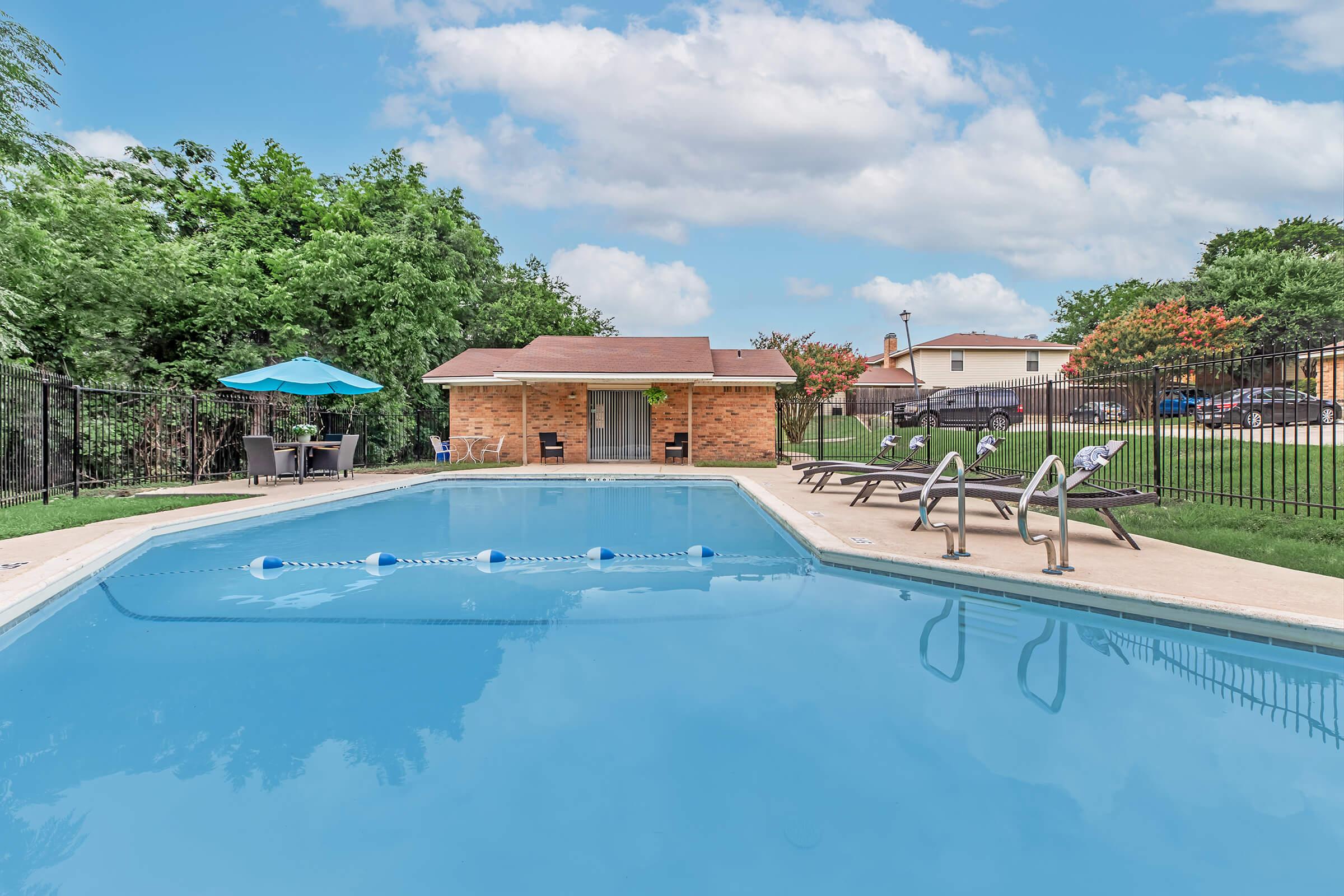 A clear blue swimming pool surrounded by green trees and a fenced area. Lounge chairs are positioned on the pool deck, and a small shaded seating area with an umbrella is visible. A brick building is in the background, with a lawn and further houses beyond the fence. The sky is partly cloudy.